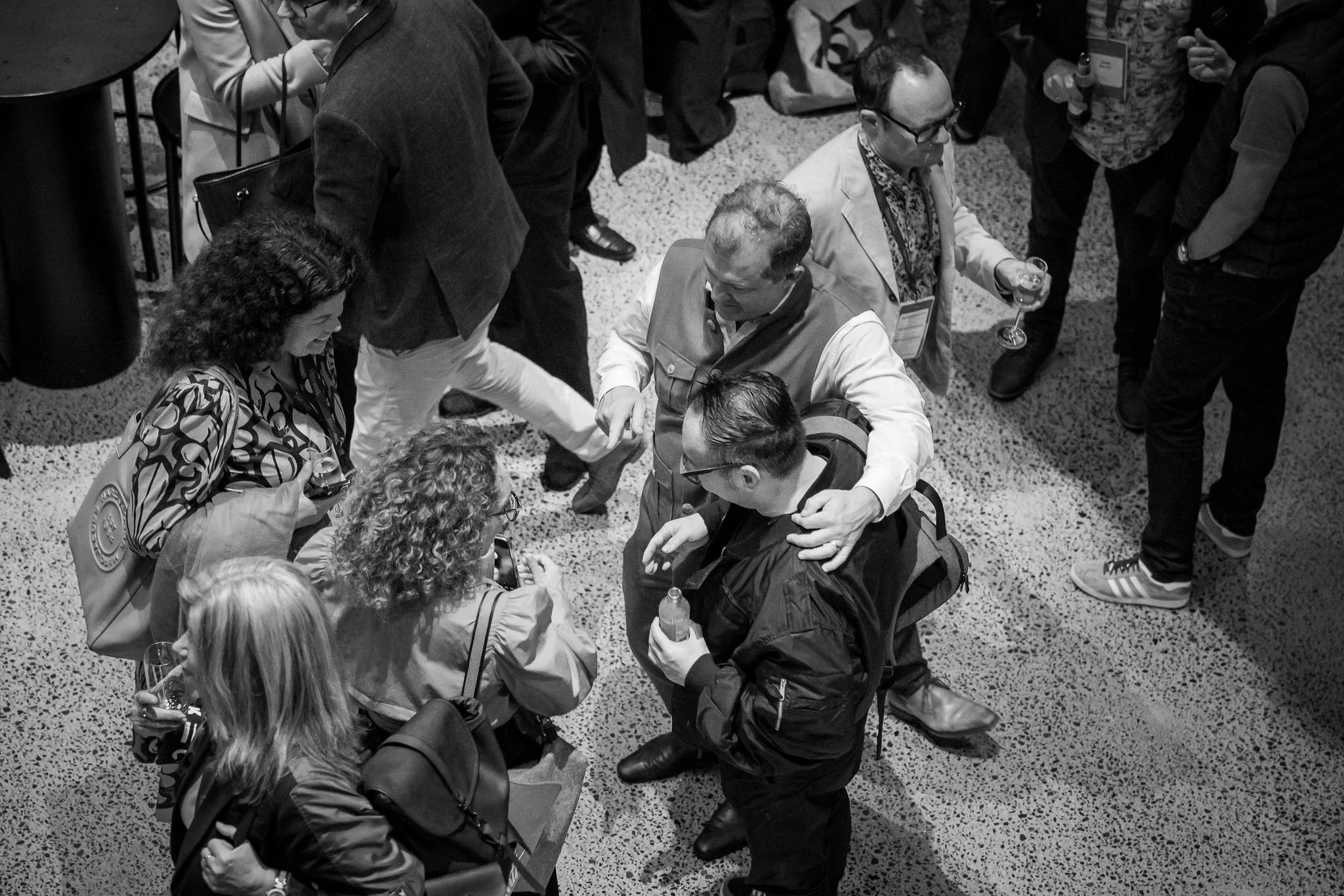 Black-and-white photo of a crowded indoor event with people engaging in conversations, some with drinks. A woman with curly hair and a patterned blouse smiles, holding a glass. A man with glasses and a patterned shirt holds a glass of wine. A man in 