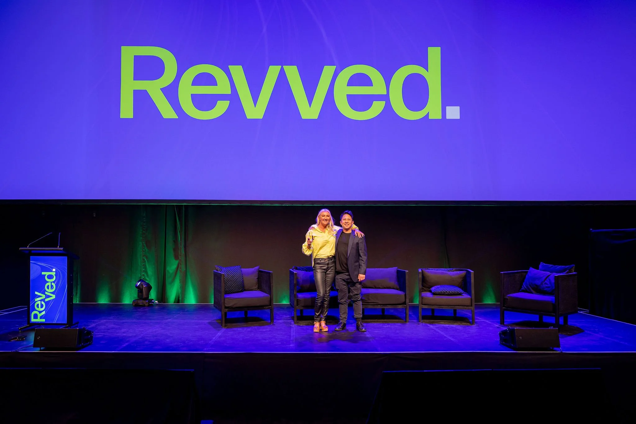 Two women standing on stage at a Reved event, smiling and posing for a photo, with a large screen behind them displaying the word 'Reved.' in green letters on a purple background.