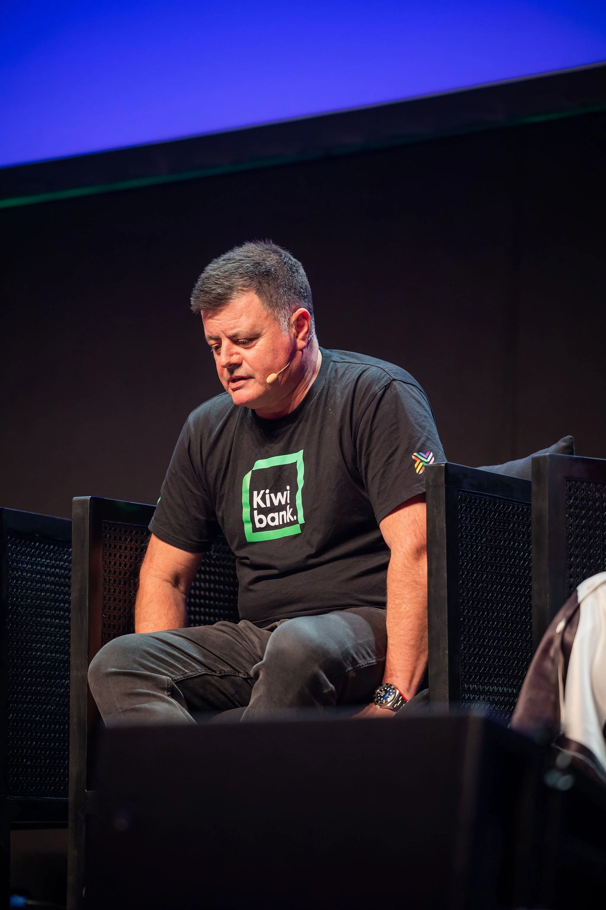 A man wearing a black T-shirt with a Kiwi Bank logo, sitting on a black chair with a distressed expression, in a dark room with a stage lighting effect.