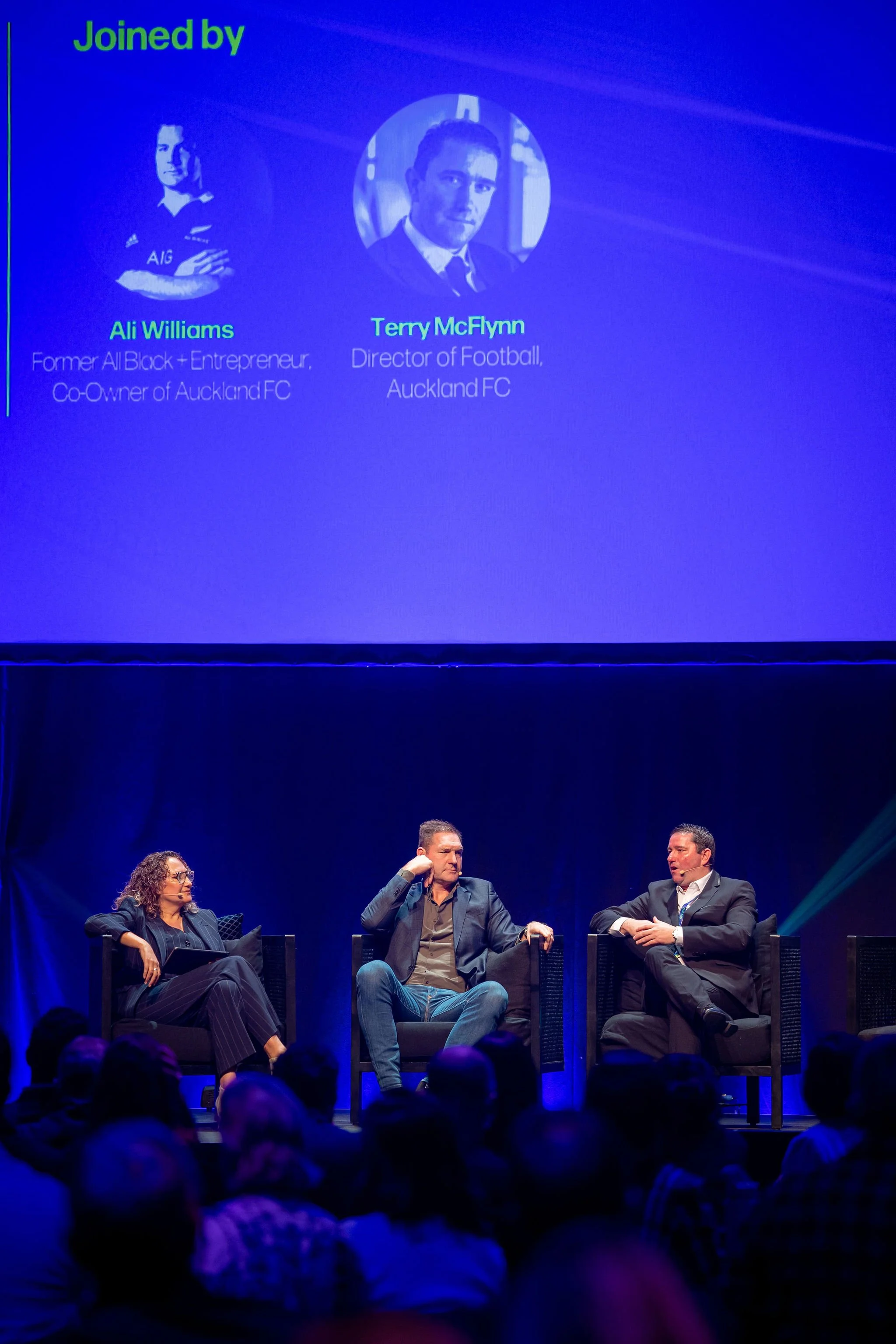 Three people sitting on chairs on stage during a panel discussion with a blue backdrop displaying names and titles. The panel includes a woman on the left, a man in the middle, and another man on the right. Audience members are visible in the foregro