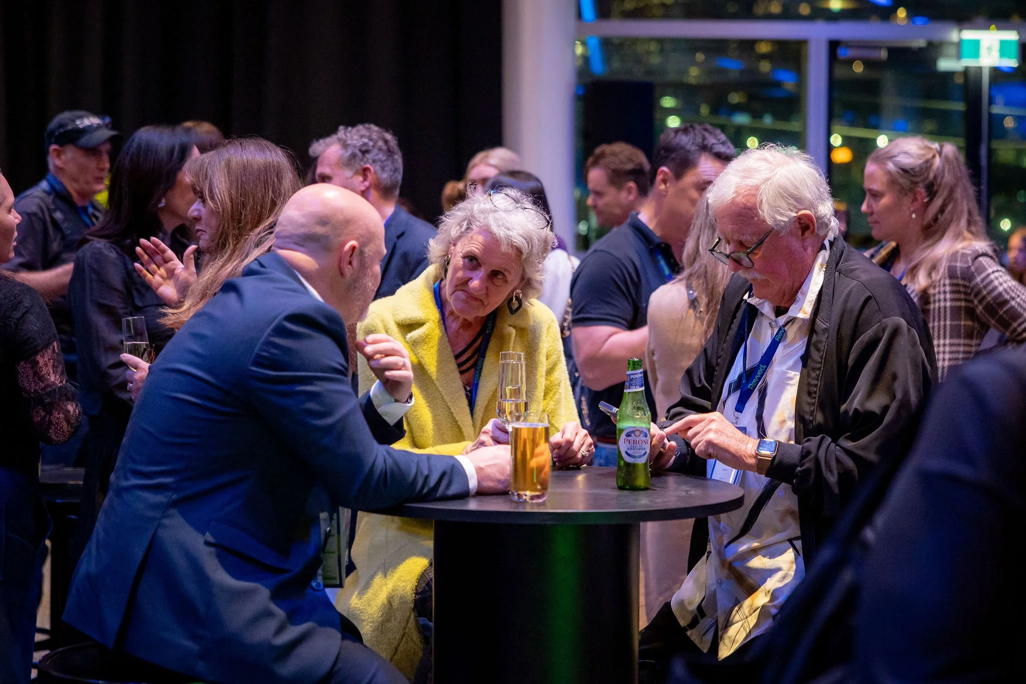 A group of people at a social gathering, some engaged in conversation, around a bar table with drinks, in an indoor setting with large windows and city lights visible outside.