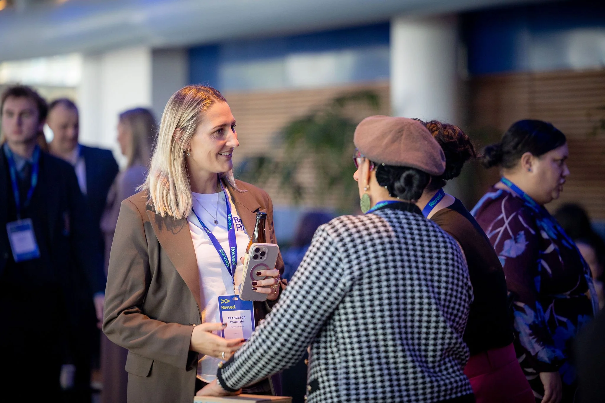 A woman with blonde hair wearing a beige blazer and white shirt holding a smartphone, engaging in conversation with another woman wearing glasses and a checkered jacket, at a professional event with other attendees in the background.