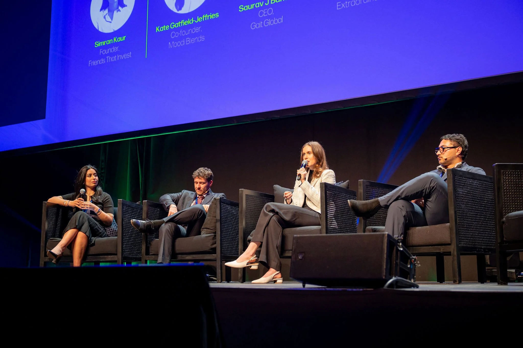 A panel discussion at a conference with five people seated on a stage, including three women and two men. One woman is speaking into a microphone. A large digital screen behind them displays panelists' names and titles.