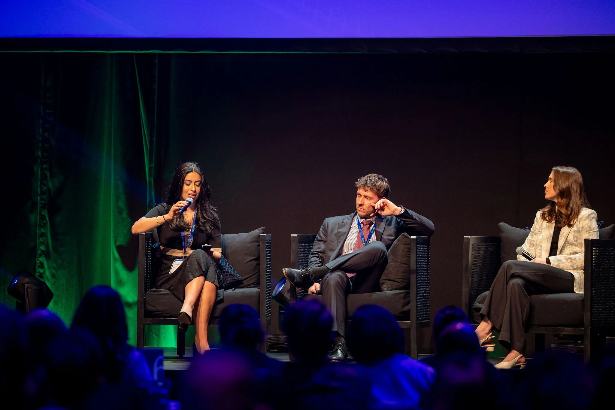 Three people seated on a stage during a panel discussion or conference, with a woman speaking into a microphone and two others listening, against a dark background with purple and green lighting.