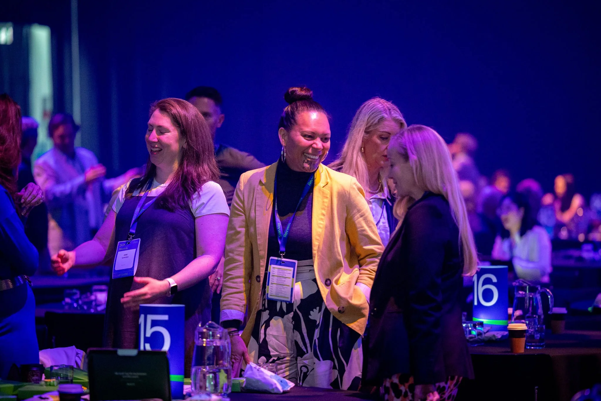 People having conversations at Revved in a dark room with blue lighting, several tables with numbered signs, and drinks on the tables.