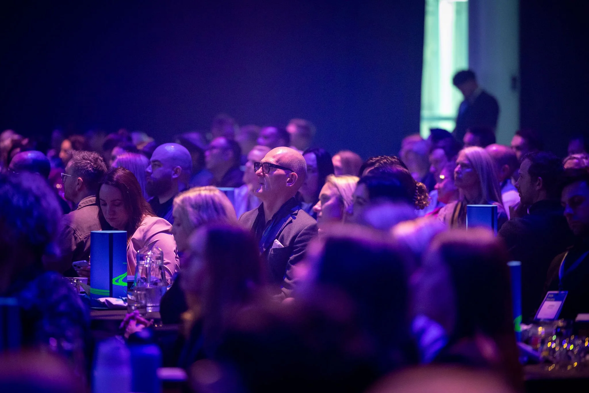 Revved conference auditorium filled with a diverse group of people sitting and listening, with some looking at their devices or talking quietly. The room is illuminated with purple and blue lighting.