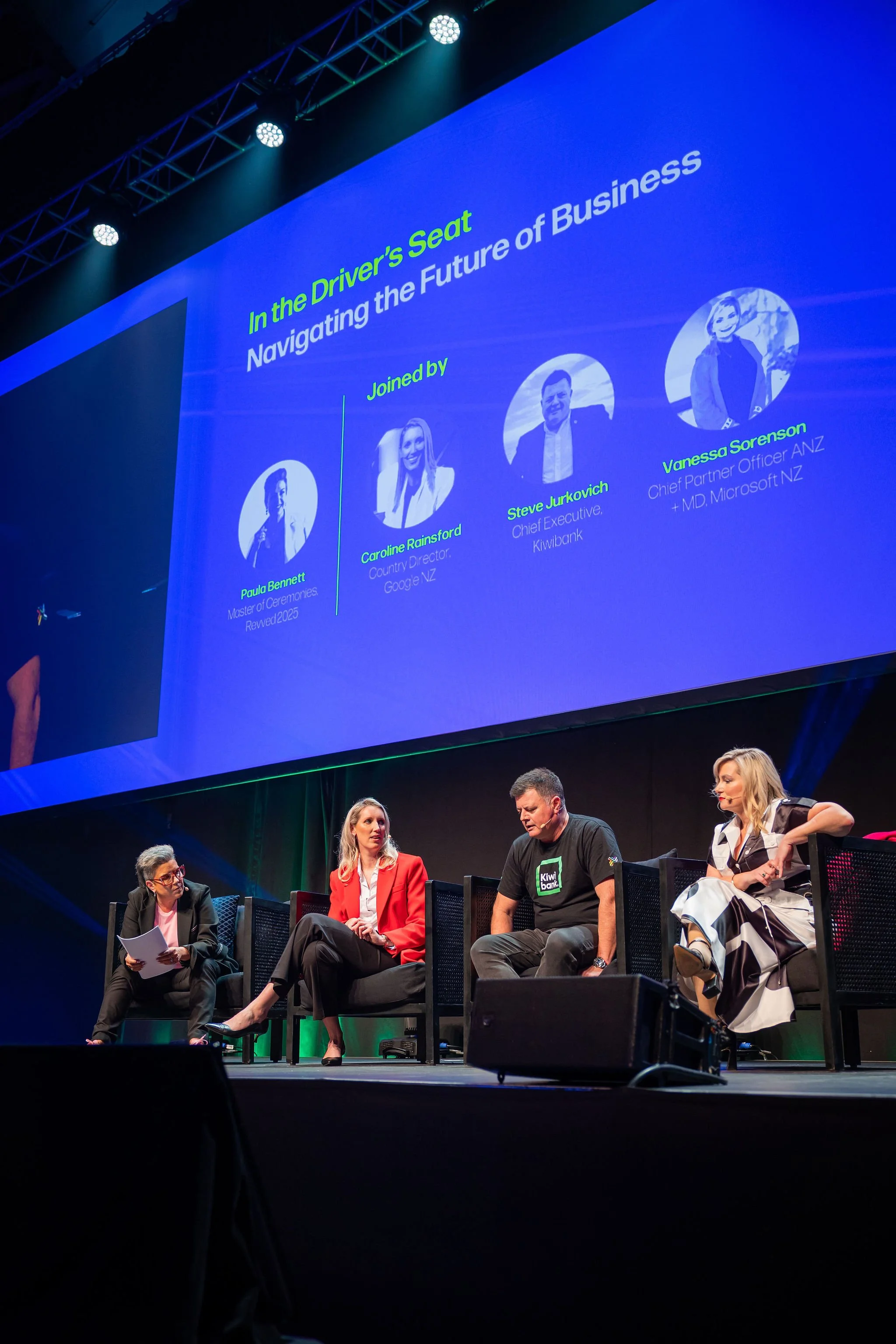 Panel discussion at a conference with four people sitting on stage, a large screen above displaying names and titles of the panelists, and a blue background. The panelists include a woman in a black suit, a woman in a red blazer, a man in a black shi