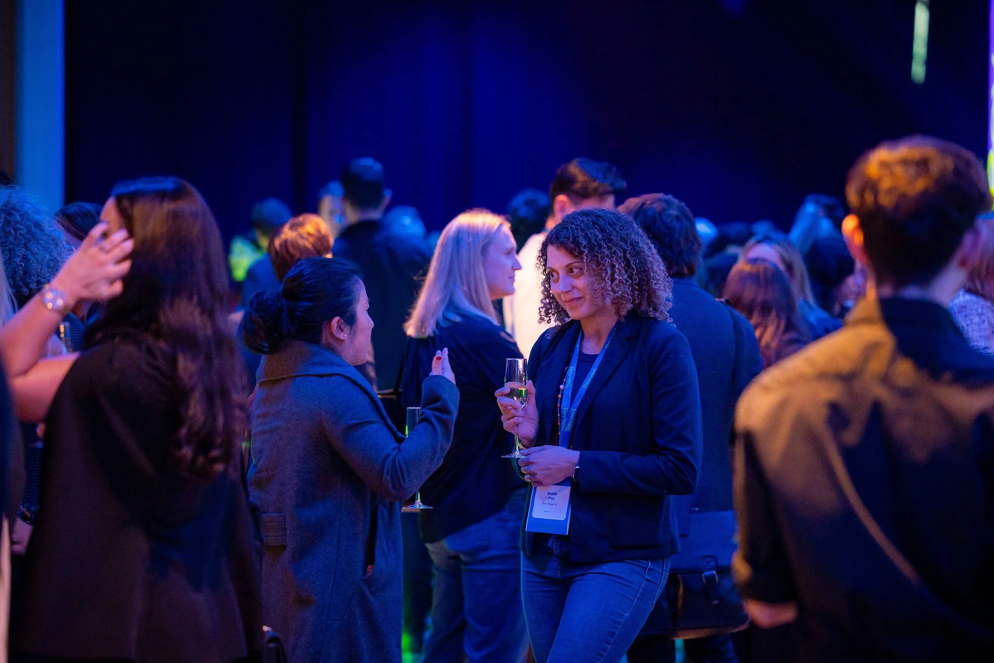 Group of people socializing at an indoor event with blue lighting, some holding glasses of wine or champagne, engaged in conversations.