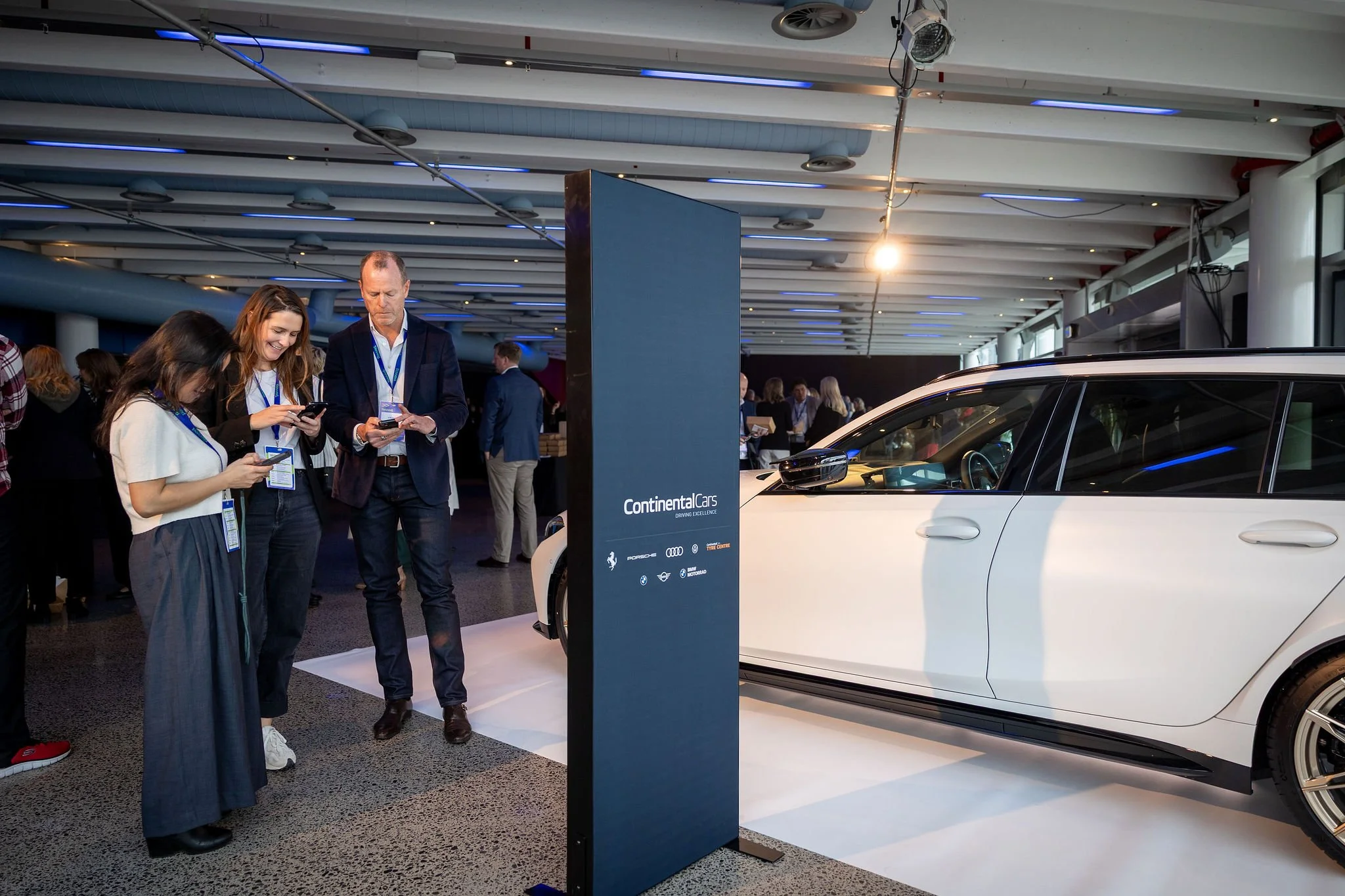 People gather around a white car at an auto show with a large vertical sign displaying the logos of Continental Cars, Porsche, Audi, and BMW, in a modern exhibition space with a high ceiling and blue lighting accents.