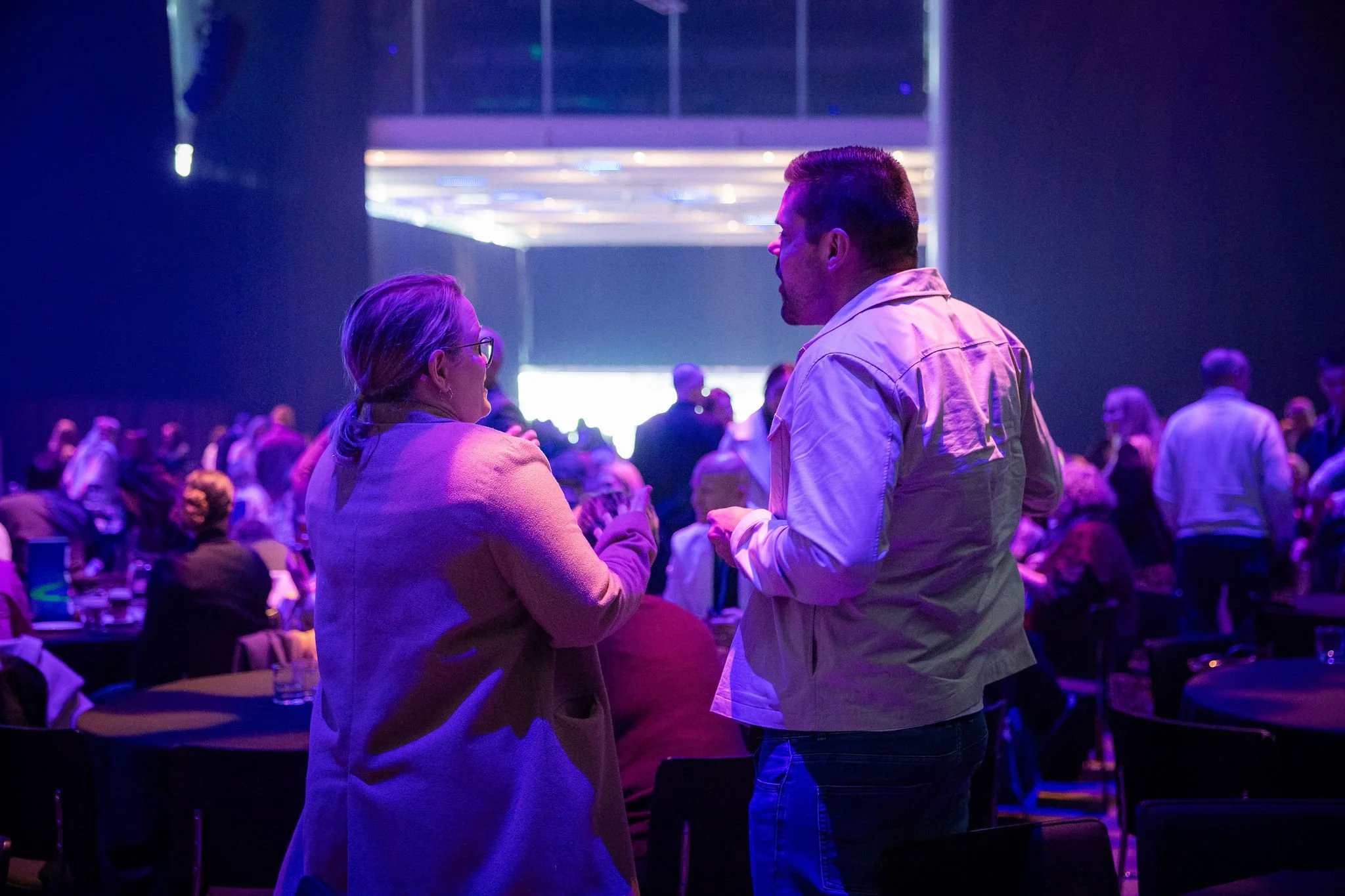 A man and woman engaged in conversation at a social event or conference, with a large crowd of people in the background illuminated by purple and blue lighting.