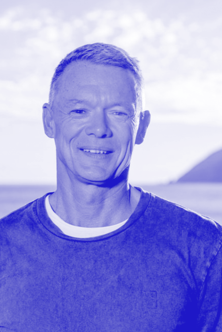 A smiling middle-aged man with short hair, wearing a dark T-shirt, standing outdoors with the sky and mountains in the background.