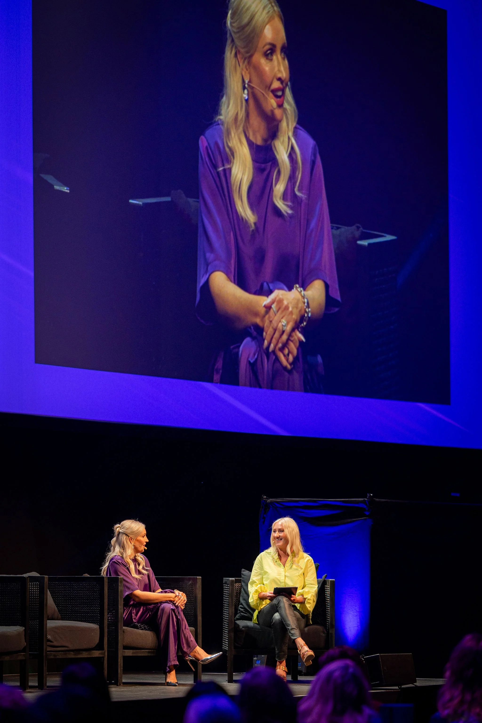 Two women sitting on stage, engaged in conversation, with an audience visible in the foreground, and a large screen projected above showing a close-up of one of the women.