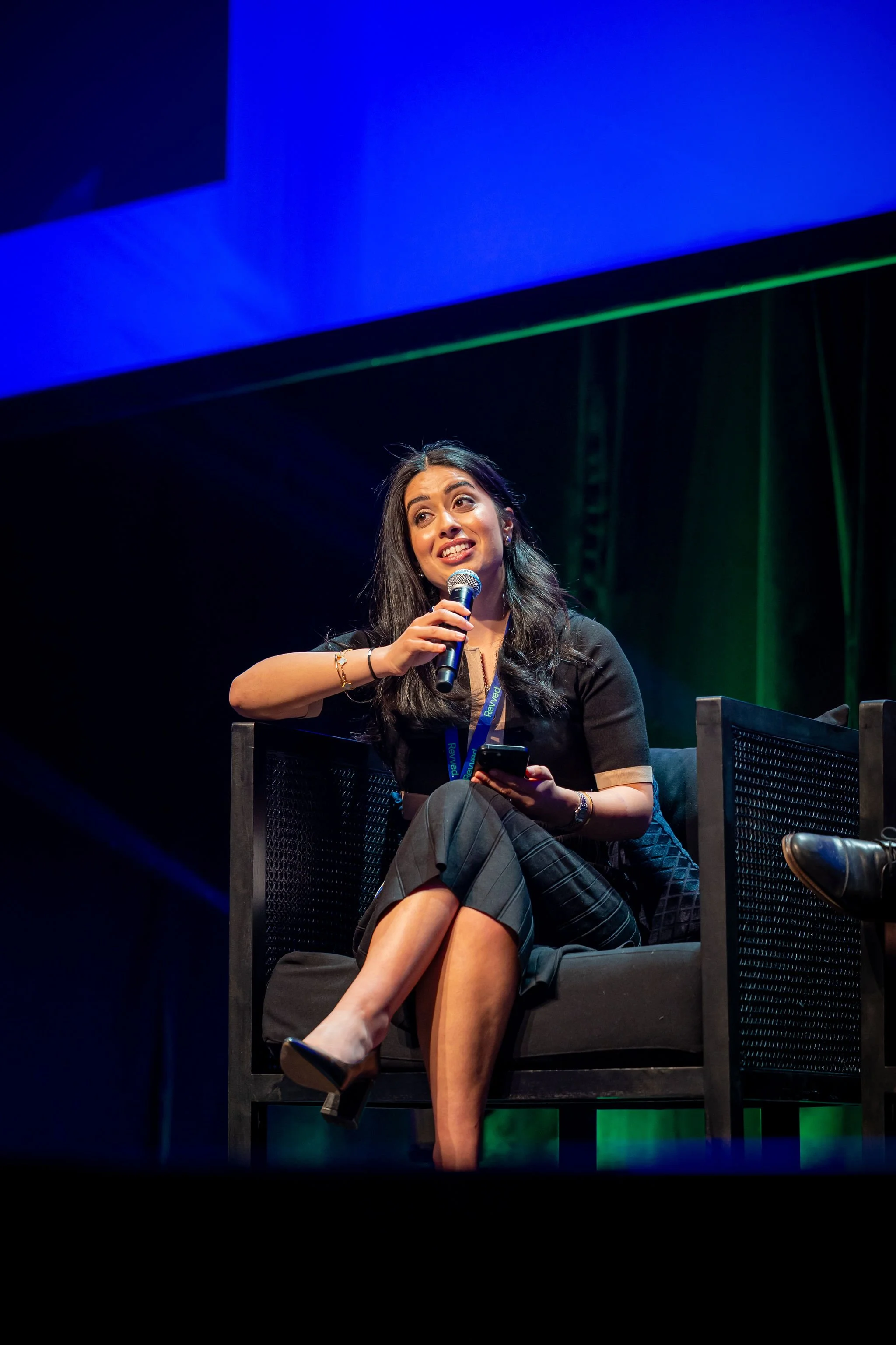 A woman sitting on a chair onstage, holding a microphone and a phone, smiling and looking upward, with a blue and black background.