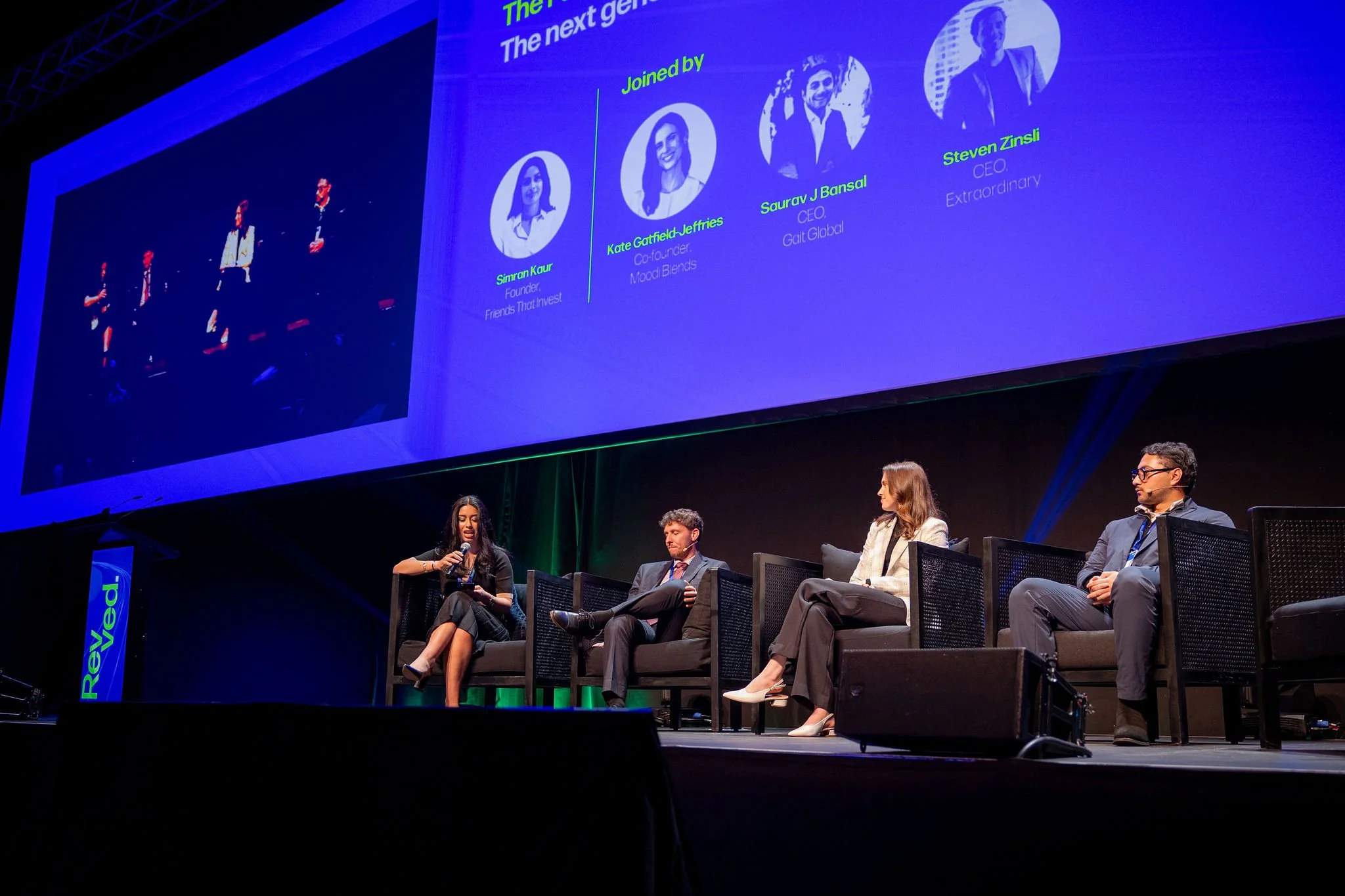 A panel discussion at a conference with five people seated on a stage and a large screen behind them displaying the names and photos of four panelists.