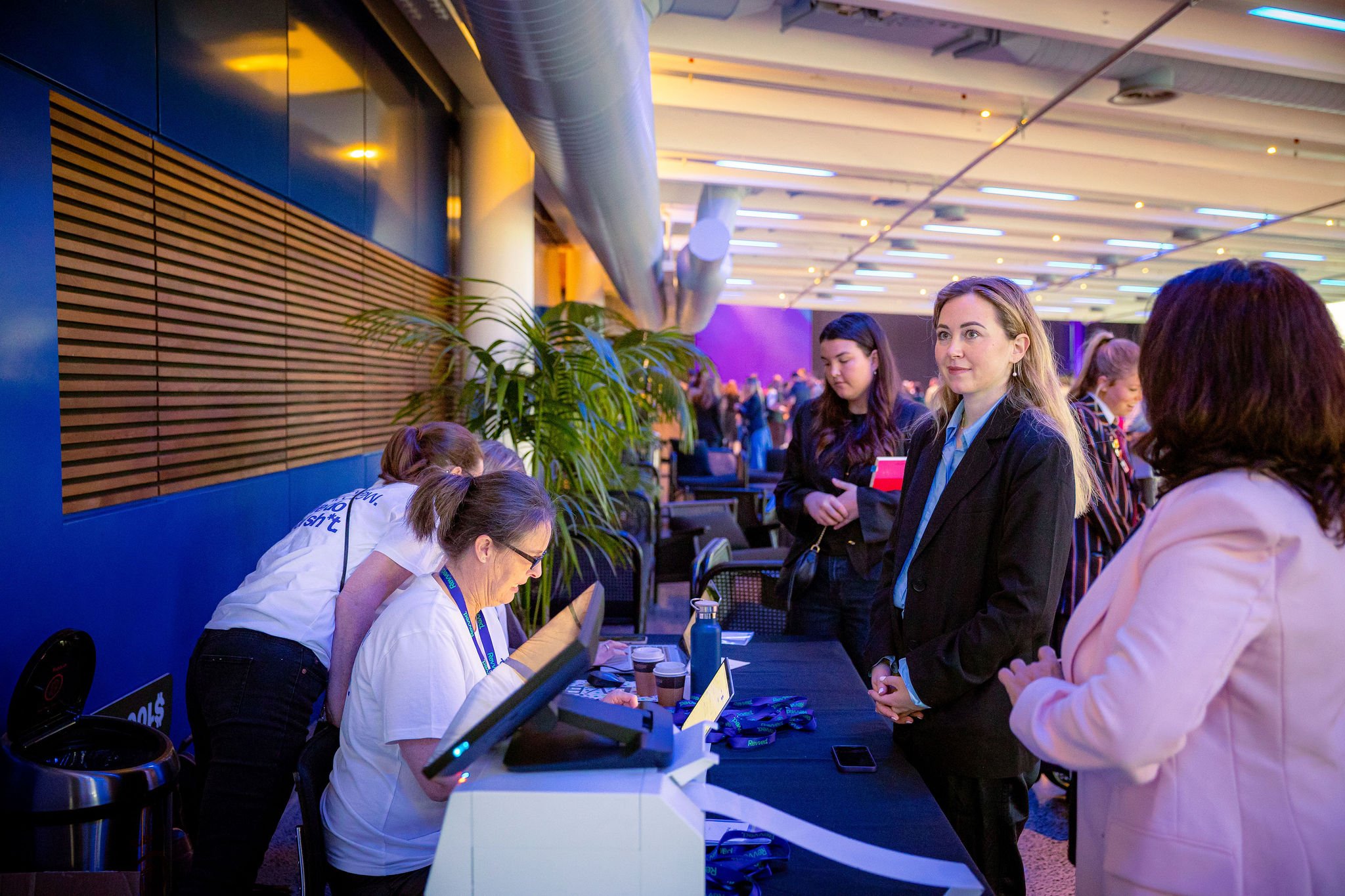 People waiting in line at a registration or check-in desk at the Revved conference, with staff behind the counter assisting attendees.