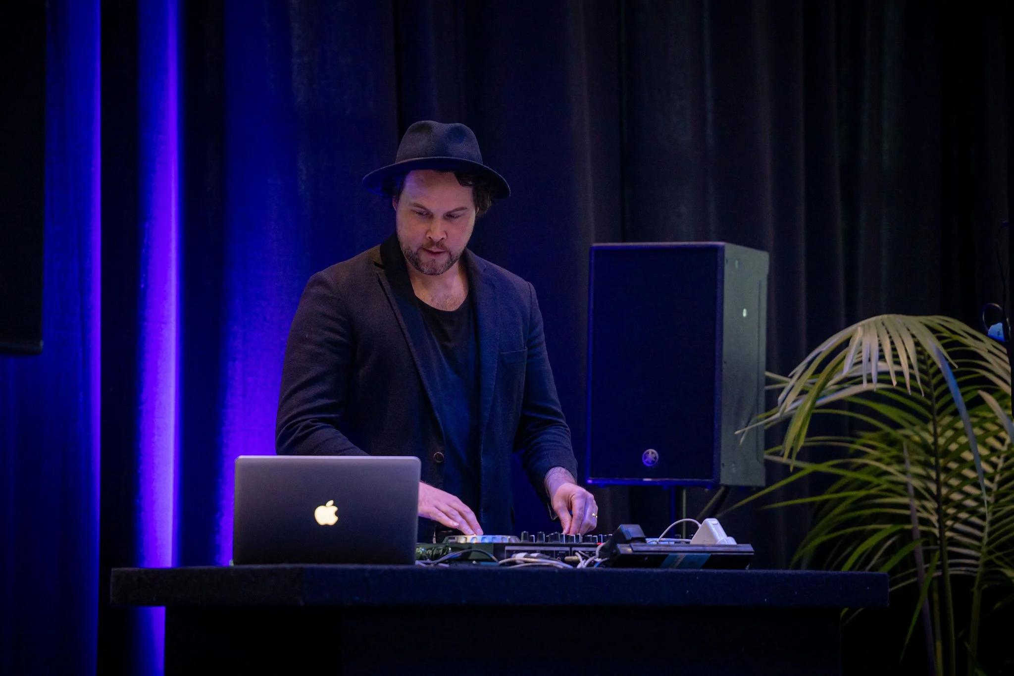 A man wearing a black hat and black clothing is working with electronic music equipment at a table, with a silver Apple MacBook laptop open in front of him, in a dark room with black curtains illuminated by blue and purple lighting.