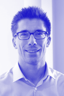 A man wearing glasses and a collared shirt, smiling, with a blurred background.