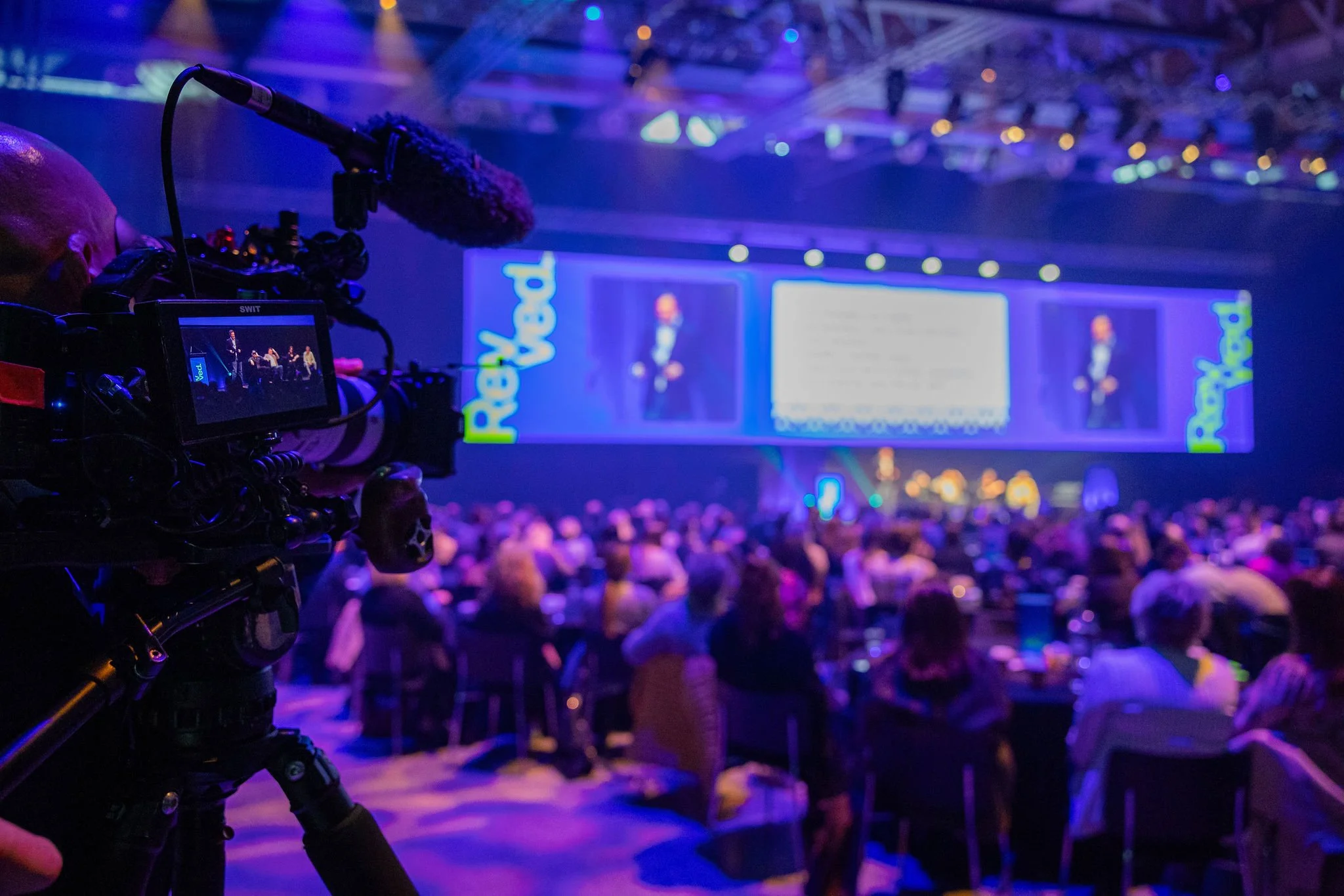 A camera operator films at the Revved conference, with a large audience. The stage has bright lights with the word 'Pod' on a large screen, and a speaker is giving a presentation.