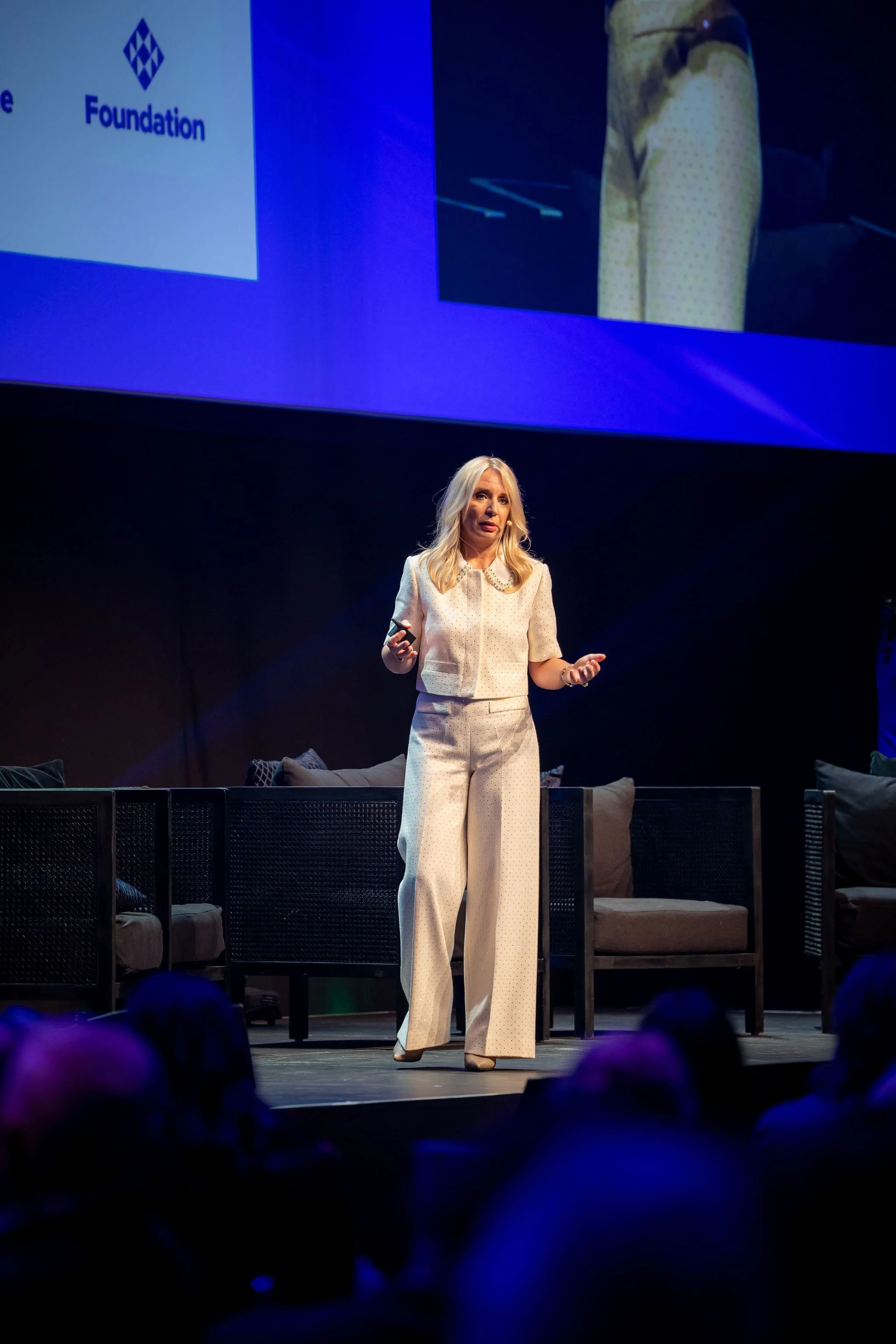 Woman in a cream-colored pantsuit giving a presentation on stage with a large screen behind her, showing a logo and text.