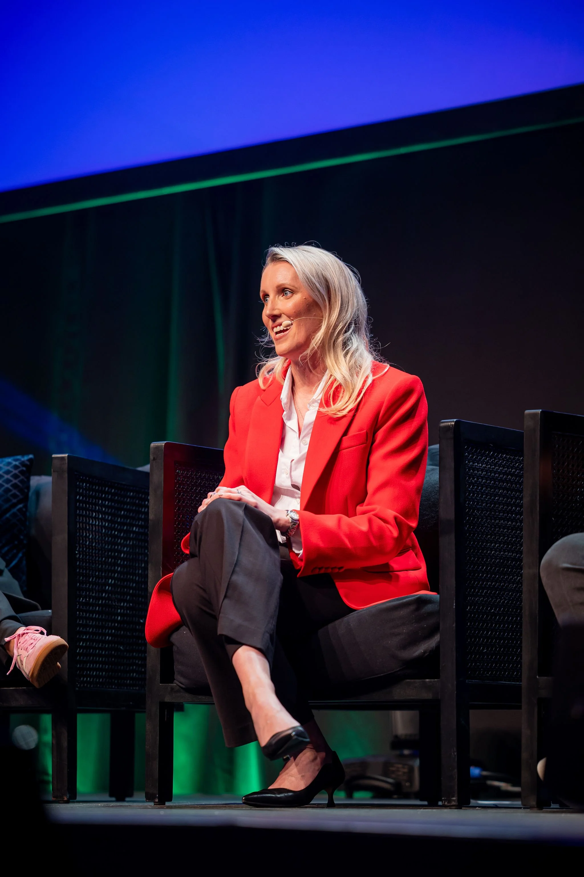 A woman with gray hair wearing a red blazer, white shirt, and black skirt, sitting on a stage in front of a black background, speaking into a headset microphone.