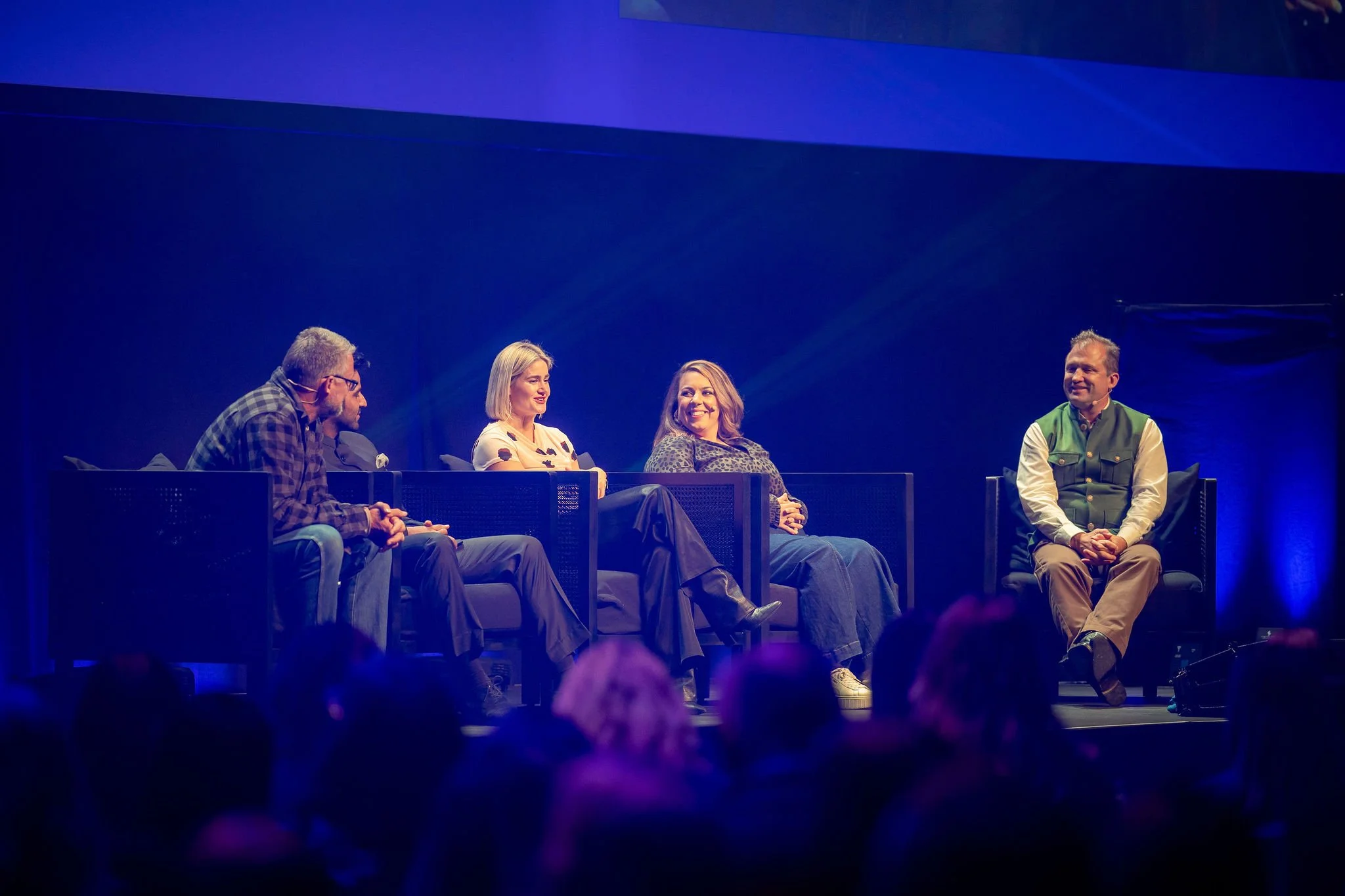 Five panelists on a stage at Revved under blue lighting, engaged in a panel discussion or conversation, with the audience visible at the bottom of the image.