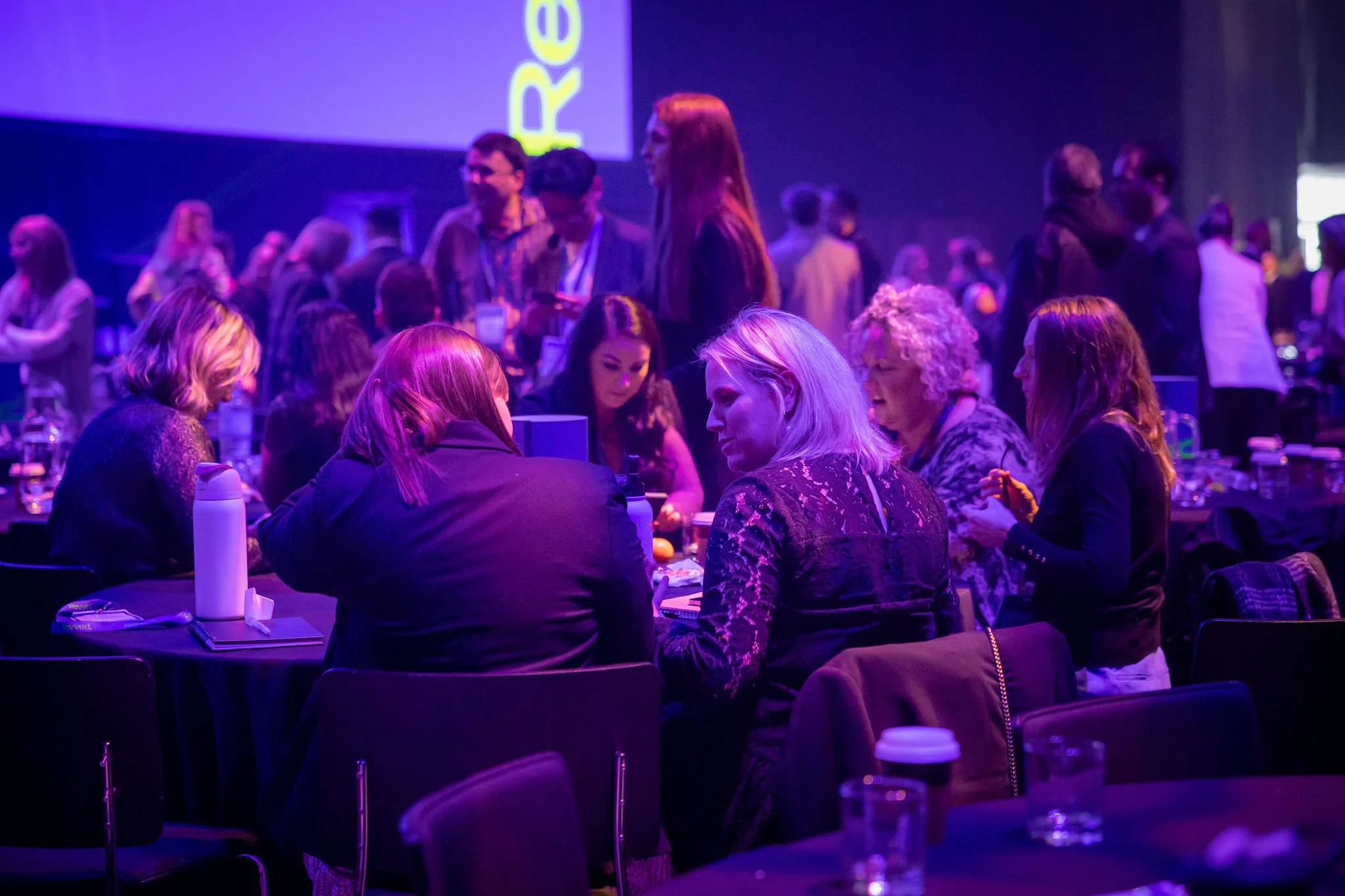People seated at tables in a dimly lit conference or event space with purple lighting, engaged in conversations.