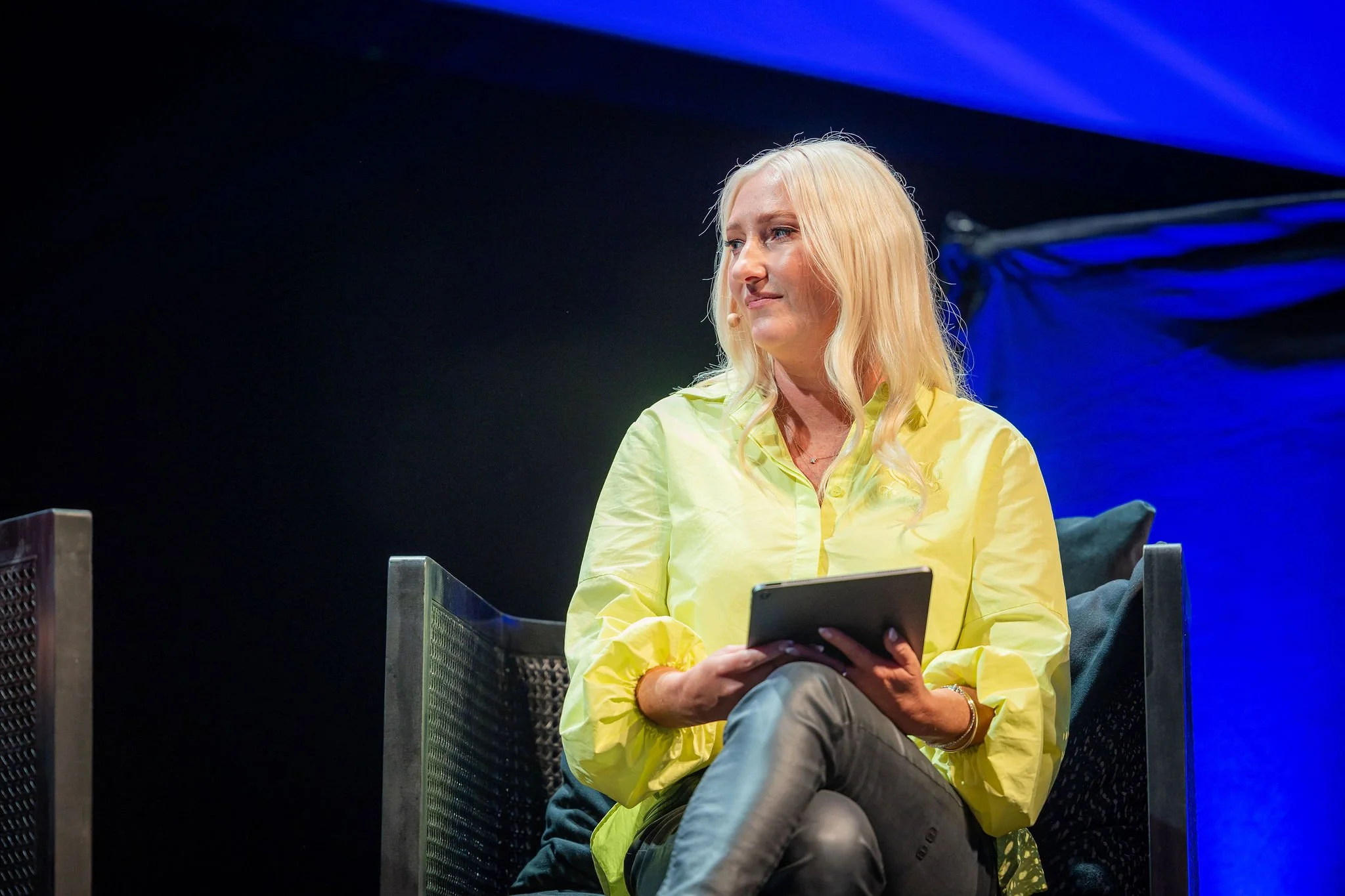 A blonde woman sitting in a dark environment, holding a smartphone, wearing a yellow jacket and gray pants.