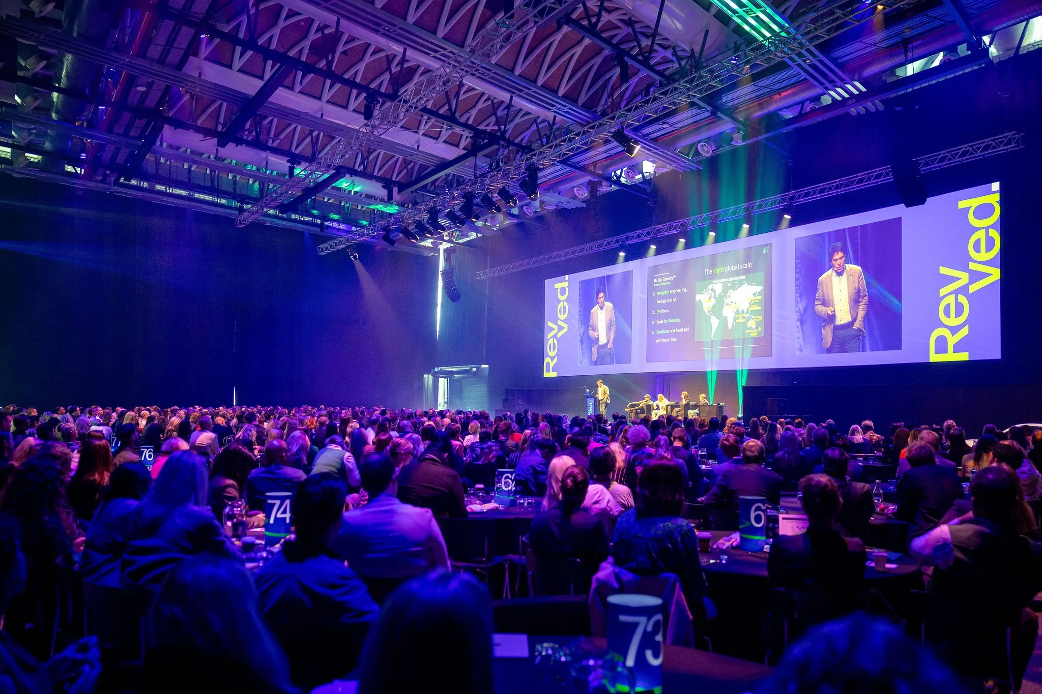 A large conference hall filled with attendees seated at round tables, facing a stage where a speaker is giving a presentation. The stage features a large screen displaying slides, and the hall is illuminated with colorful lights and rigging overhead.
