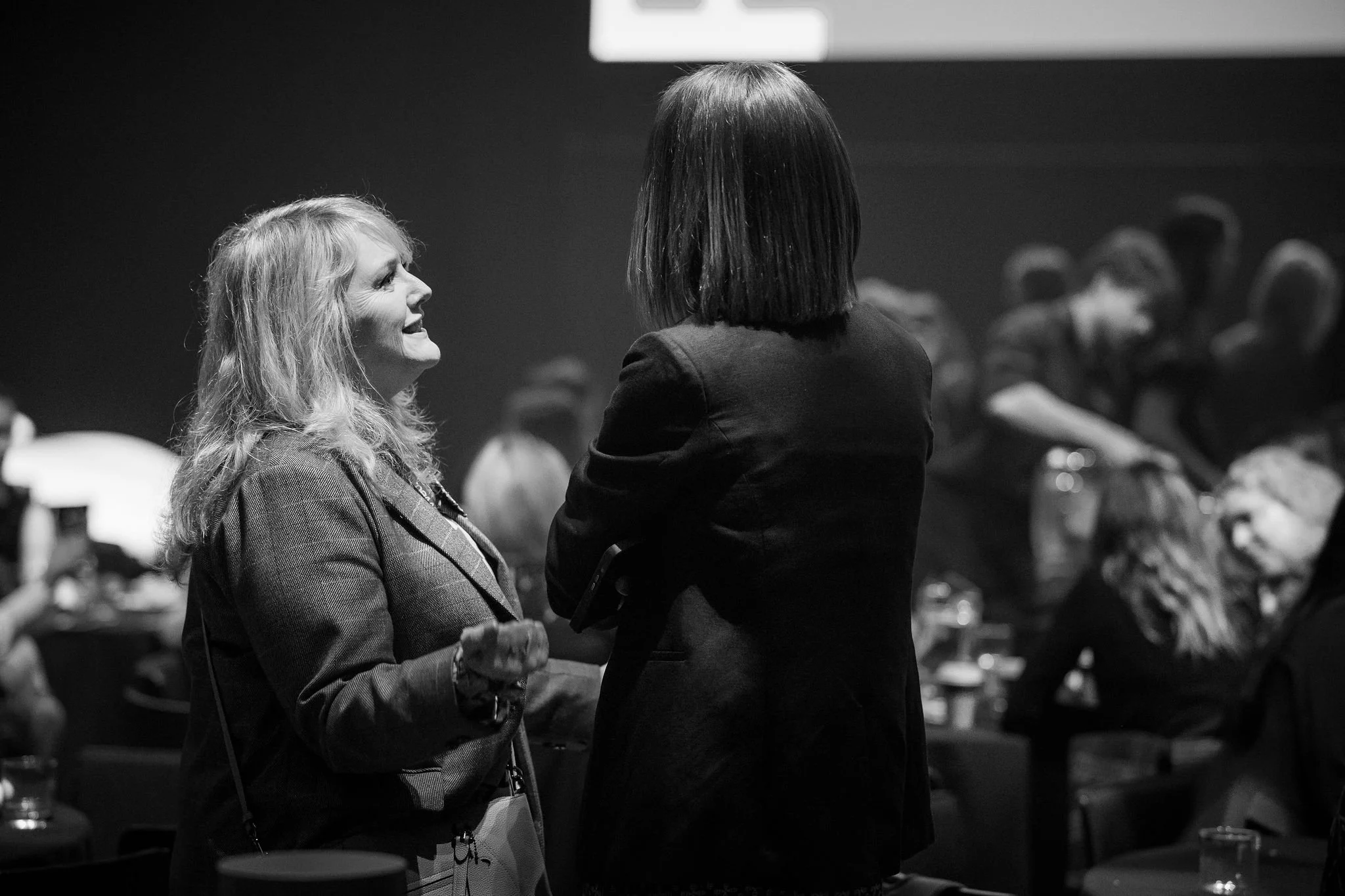 Two women are engaged in conversation, with one smiling and the other facing away, in a black-and-white setting with blurred background including a group of seated people.