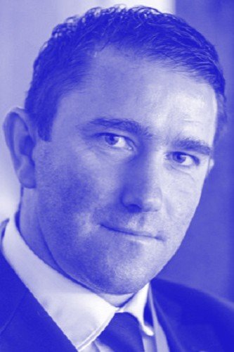 Black and white portrait of a man in a suit and tie with short, curly hair, looking into the camera.