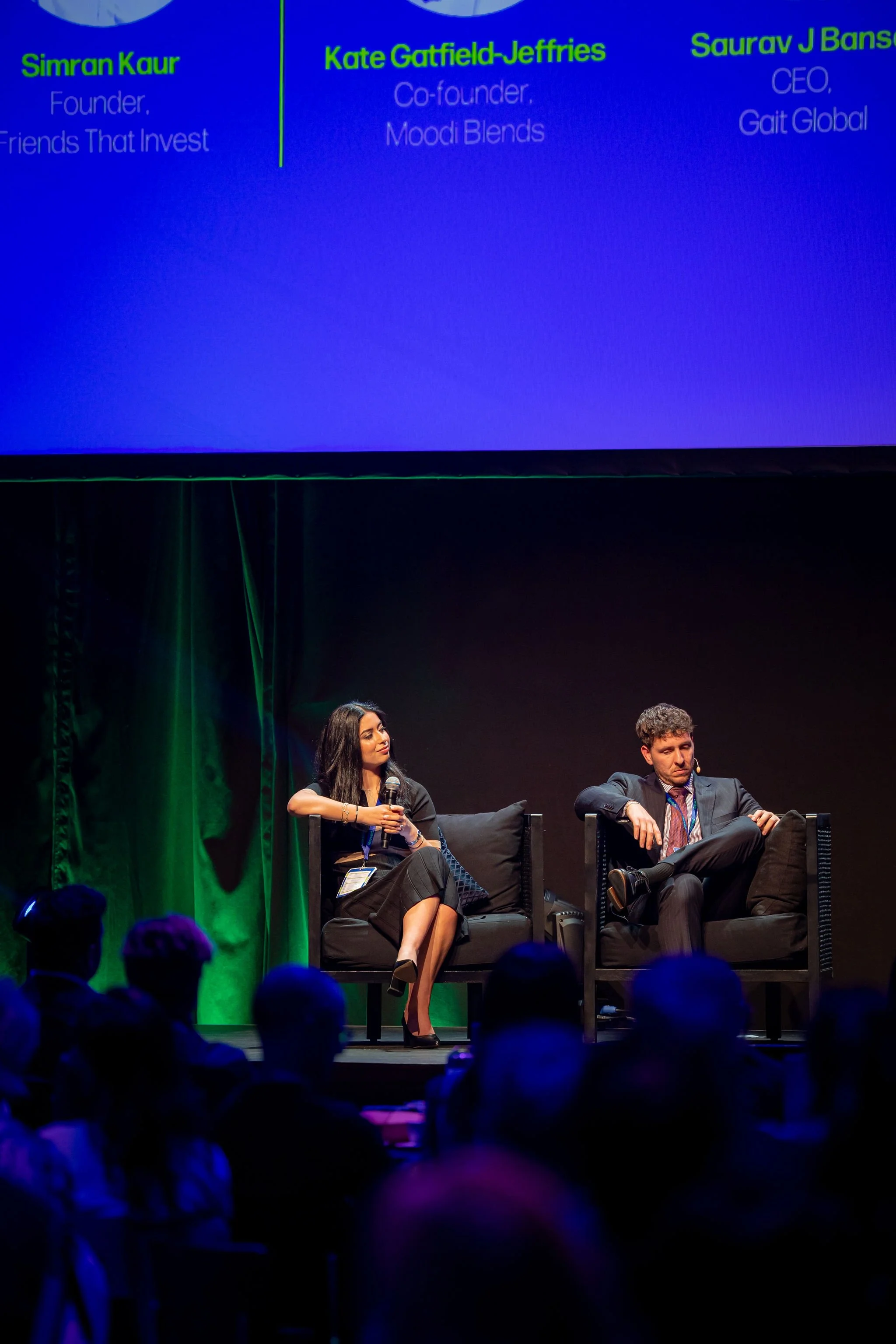 Two panelists sitting on stage at a conference, with a large blue screen behind them displaying their names and titles. The woman on the left is holding a microphone and looking to her right, dressed in a patterned skirt and blazer, with dark hair. T