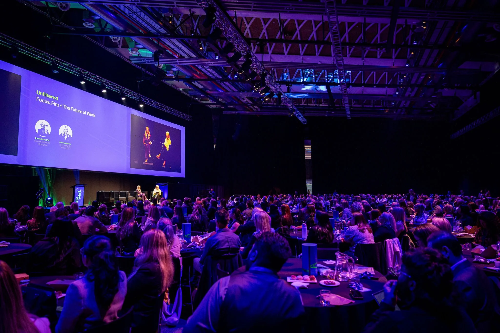 Large conference hall filled with attendees sitting at round tables, facing a stage with large screens displaying a presentation. The room is dimly lit with purple and blue lighting, and the ceiling has visible rigging and lighting equipment.