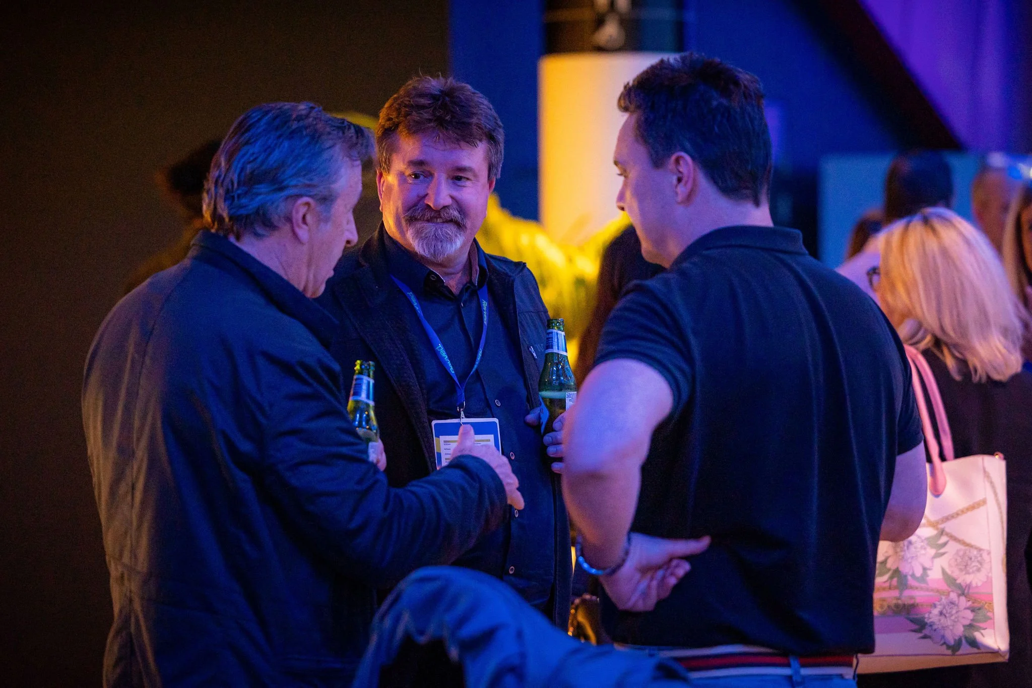 Three men are engaged in conversation at a social event, holding bottles of beer, with a background of other guests and colorful lighting.
