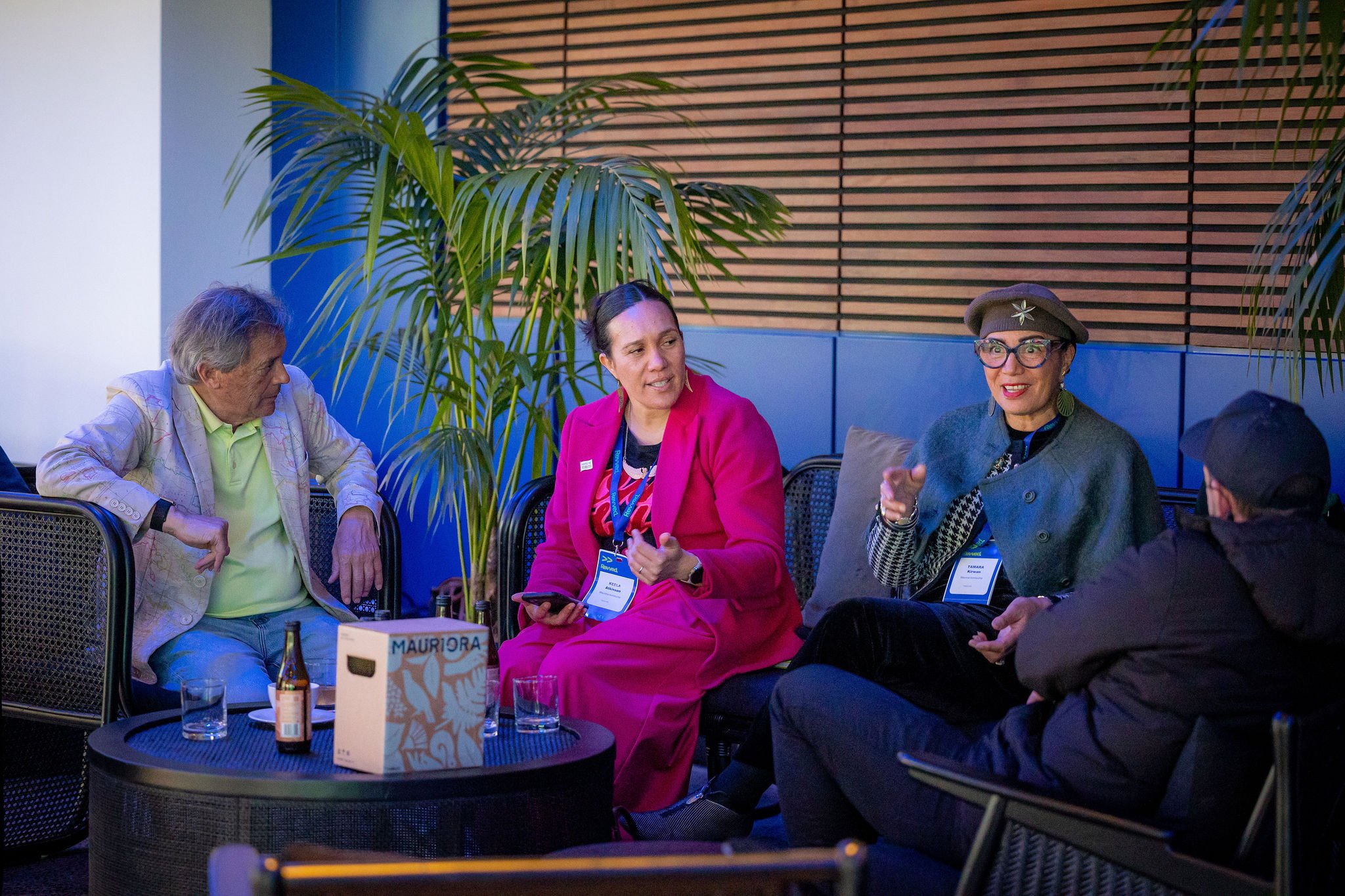 Group of four people sitting and talking at a social event. Two women and two men, some with name badges, are engaged in conversation, with drinks on the table in front of them.