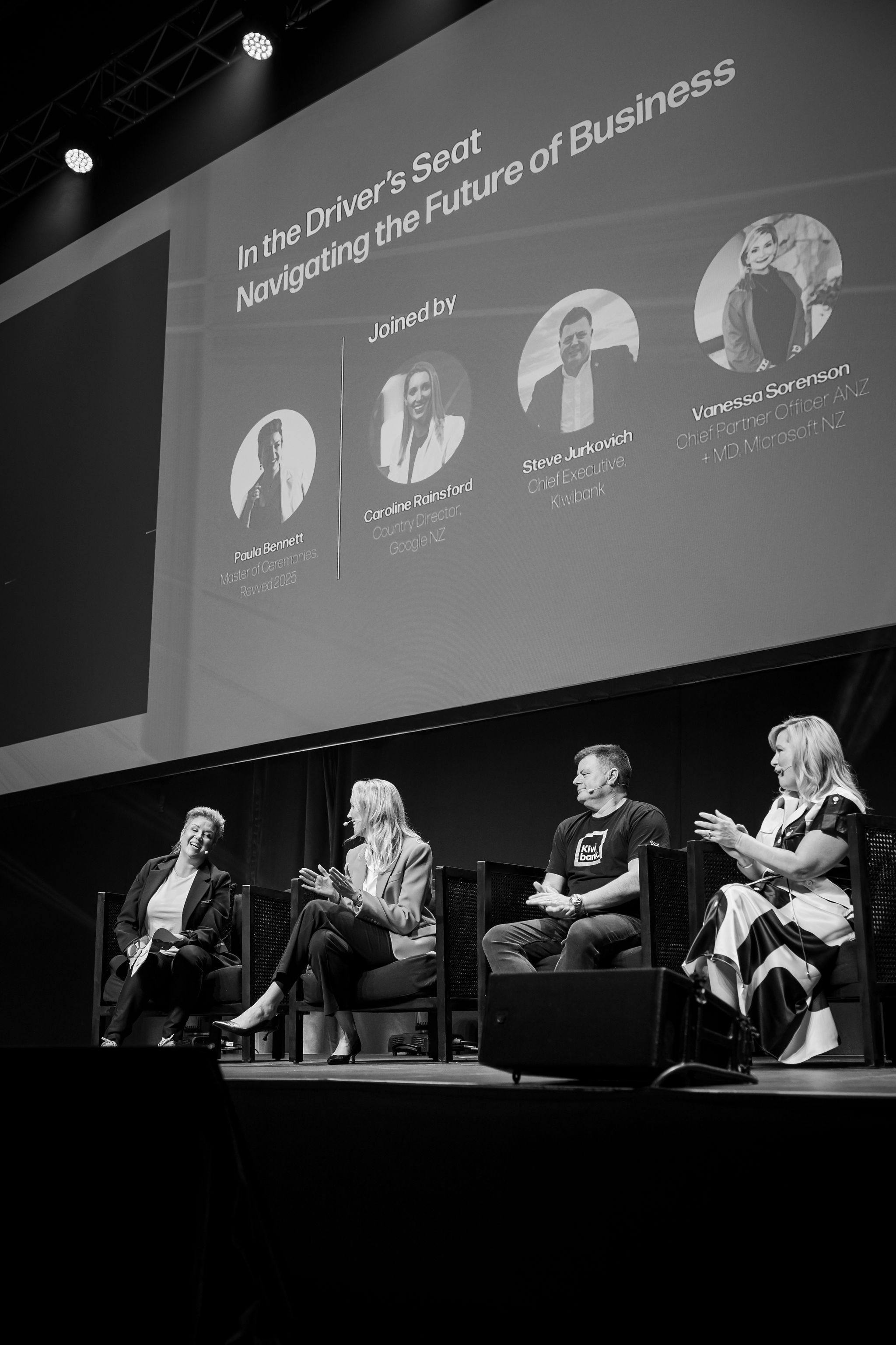 A panel discussion at a conference with five people sitting on stage in front of a large screen. The screen displays a presentation slide with photos and names of four speakers, including Caroline Rainsford, Steve Jurkovich, Vanessa Sorenson, and Pau