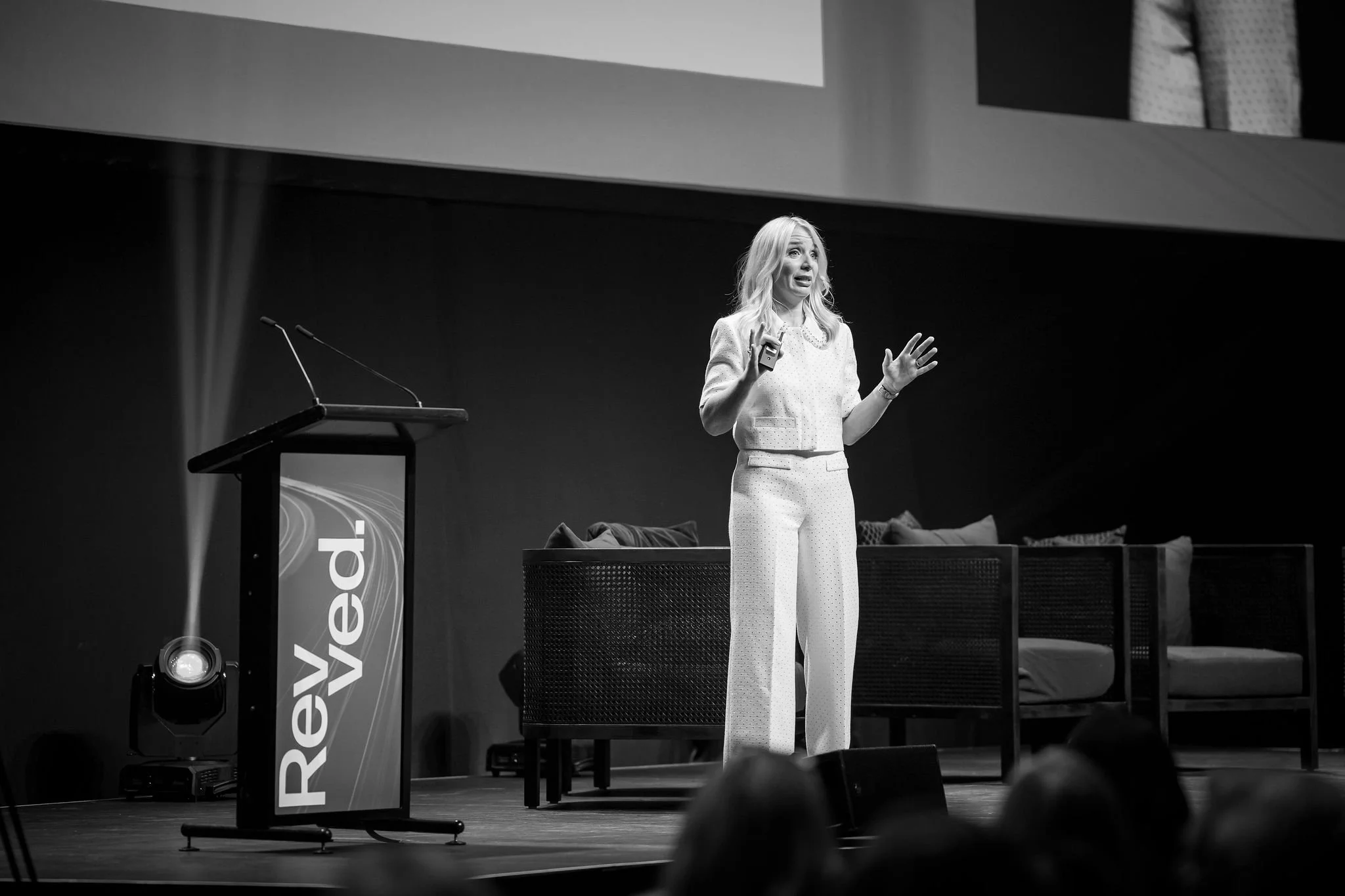 A woman with long blonde hair wearing a light-colored suit is speaking on stage at a conference or event. She appears to be gesturing with her hands while addressing an audience. There is a podium with the word 'Review' on it and a large screen above