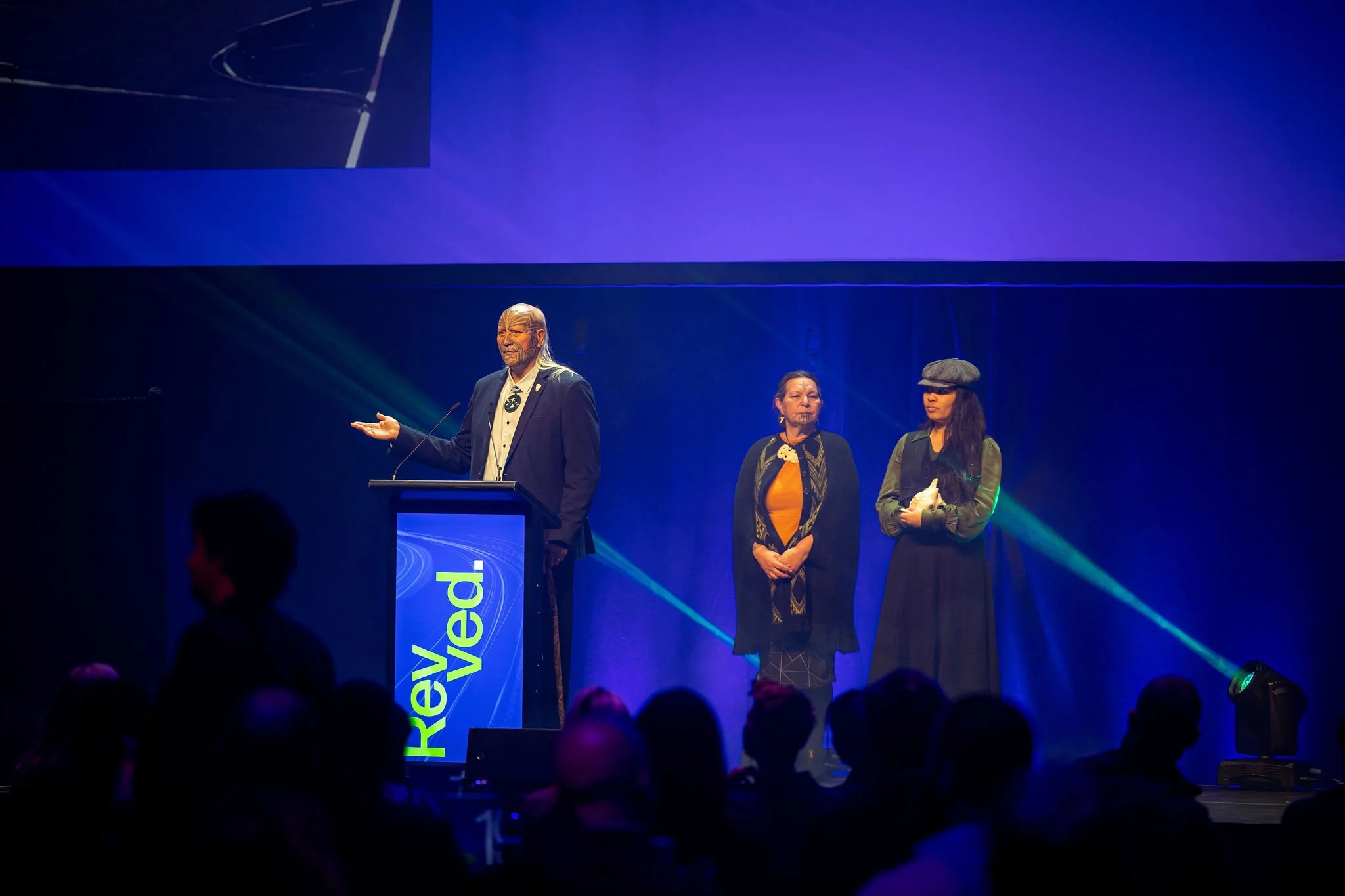 A group of three people standing on stage at Revved, with one person speaking at a podium. There is a large digital screen displaying the words 'Reved' behind them. The audience is visible in silhouette in the foreground.