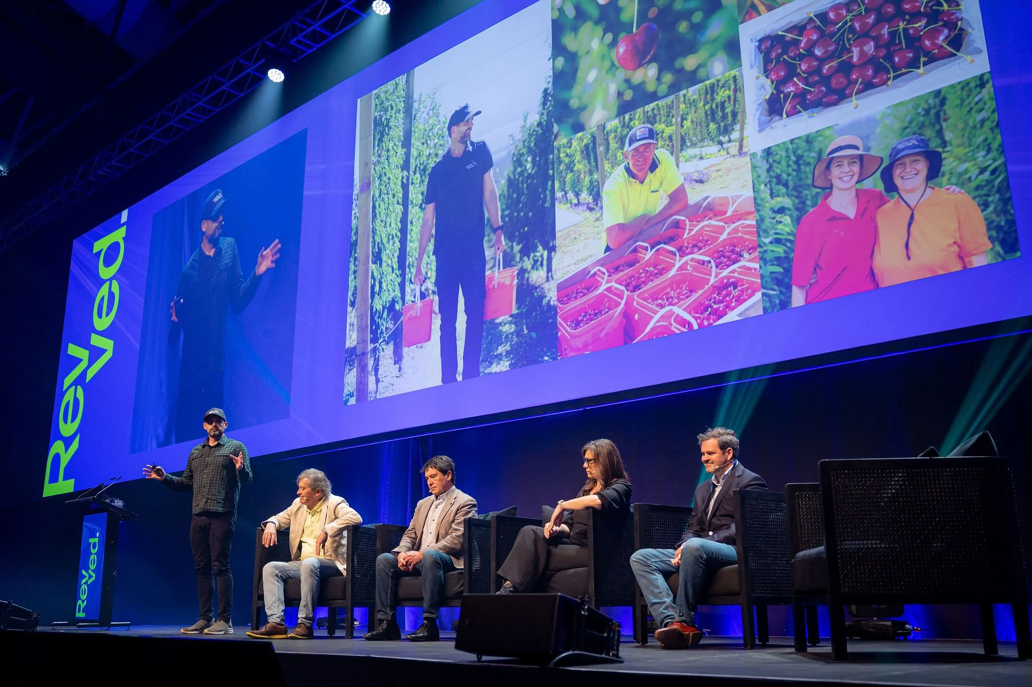 A group of people is on stage at Revved participating in a panel, with a large screen behind them displaying images of berries, cherries, and people in an orchard.