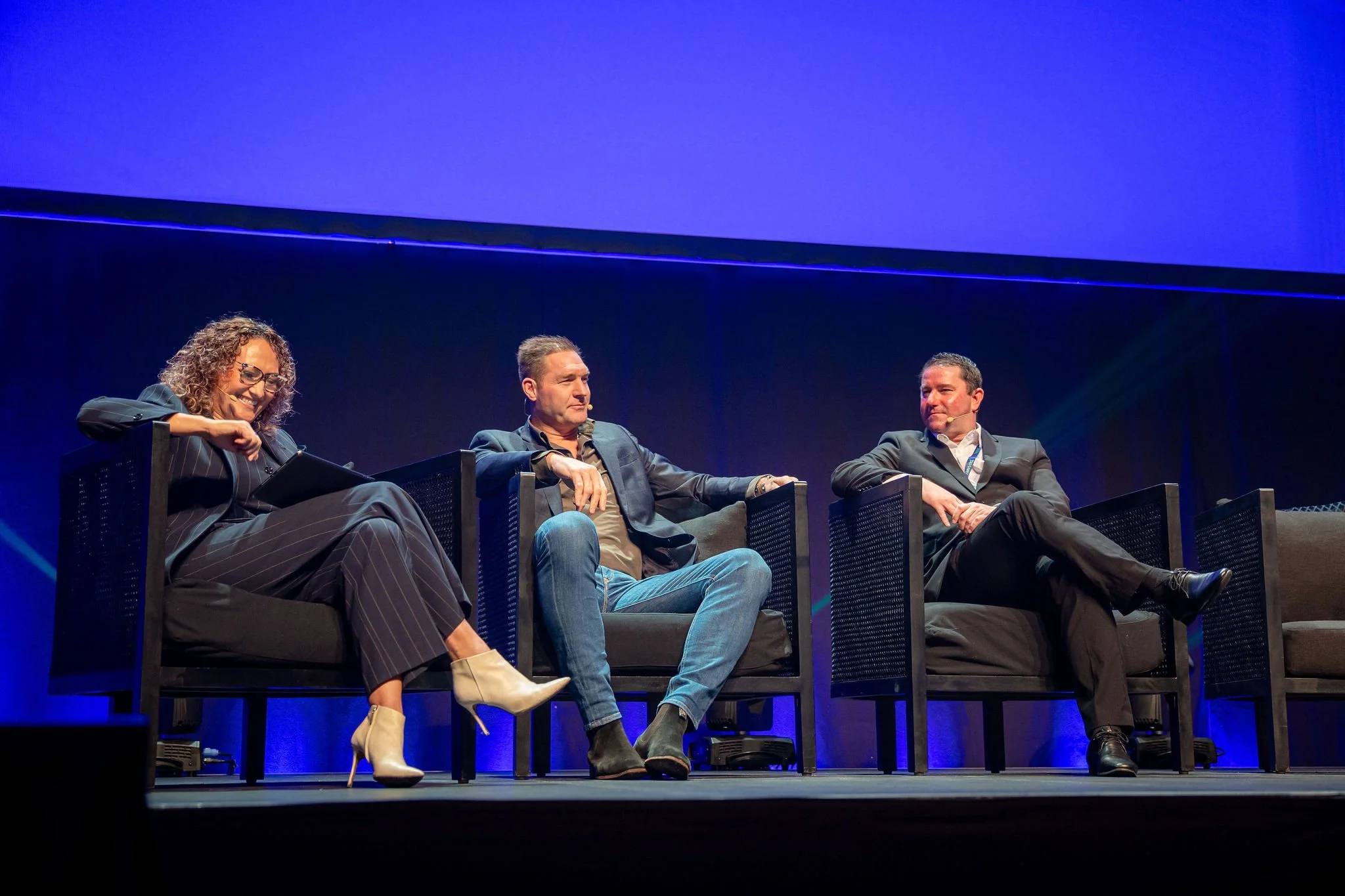 Revved panelists sitting on stage during a conference or discussion, with blue lighting and dark backdrop.
