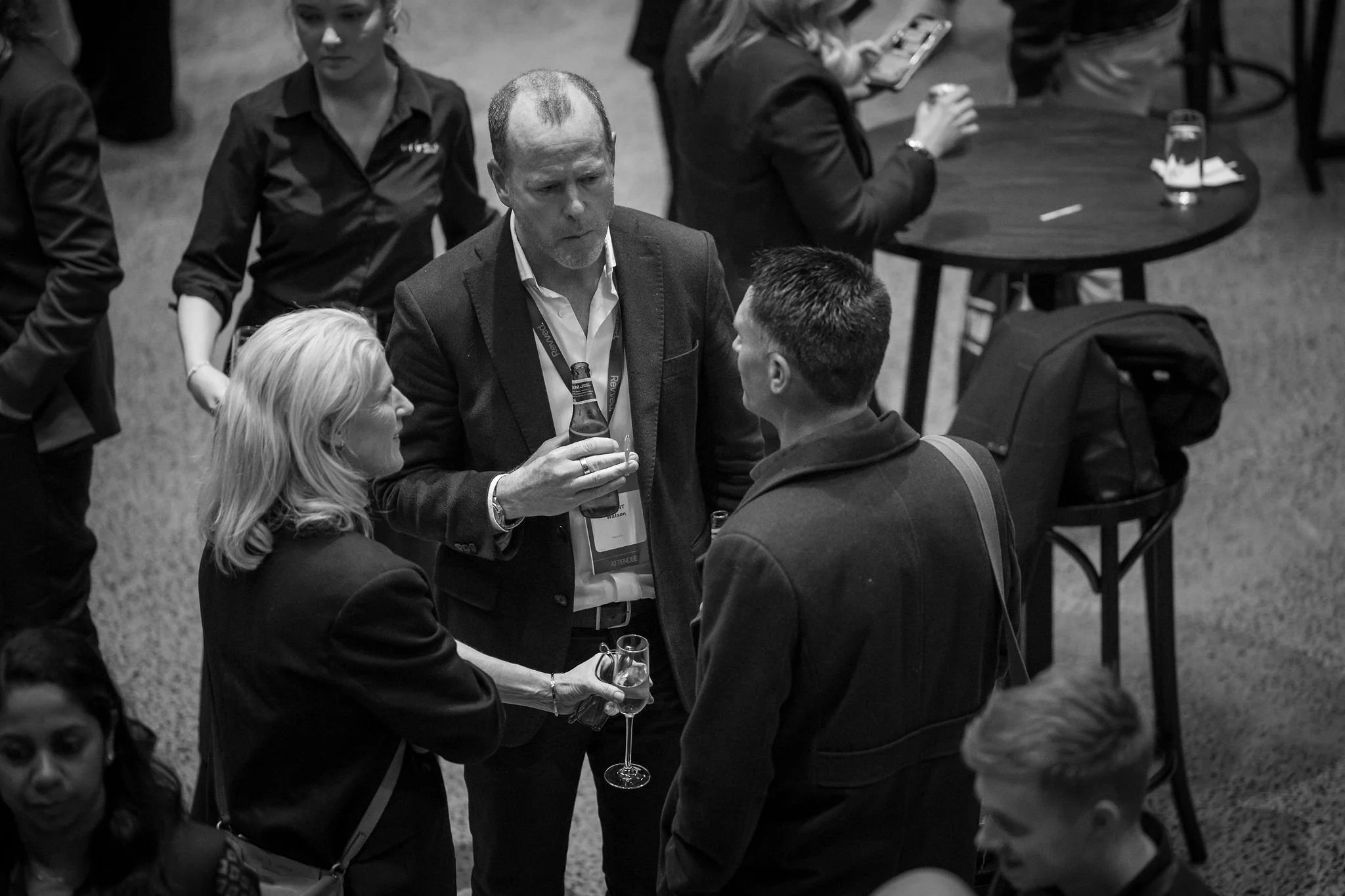 Several people at a social event, including a man with a badge and a lanyard holding a drink, engaged in conversation, with others in the background around a table with drinks.