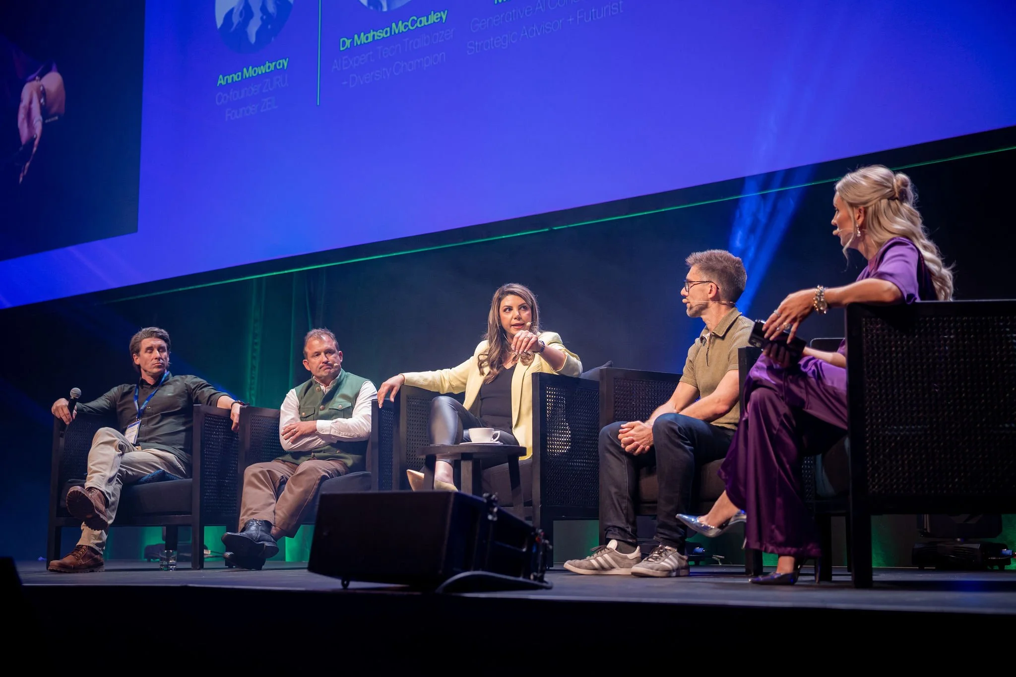 A panel discussion with six people seated on a stage, engaging in conversation. Behind them, a large screen displays the names Anna Mowbray, Dr. Mahsa McCauley, and other panelists, with blue lighting and professional setting.