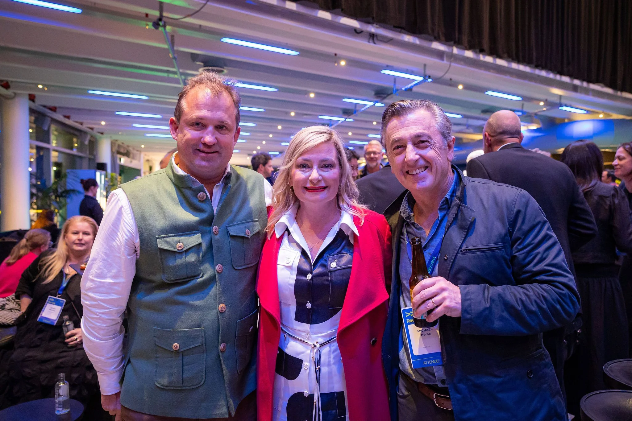 Three adults standing together at an indoor event, smiling at the camera. The man on the left is wearing a green vest over a white shirt, the woman in the middle has blonde hair and is wearing a red blazer over a white and black top, while the man on