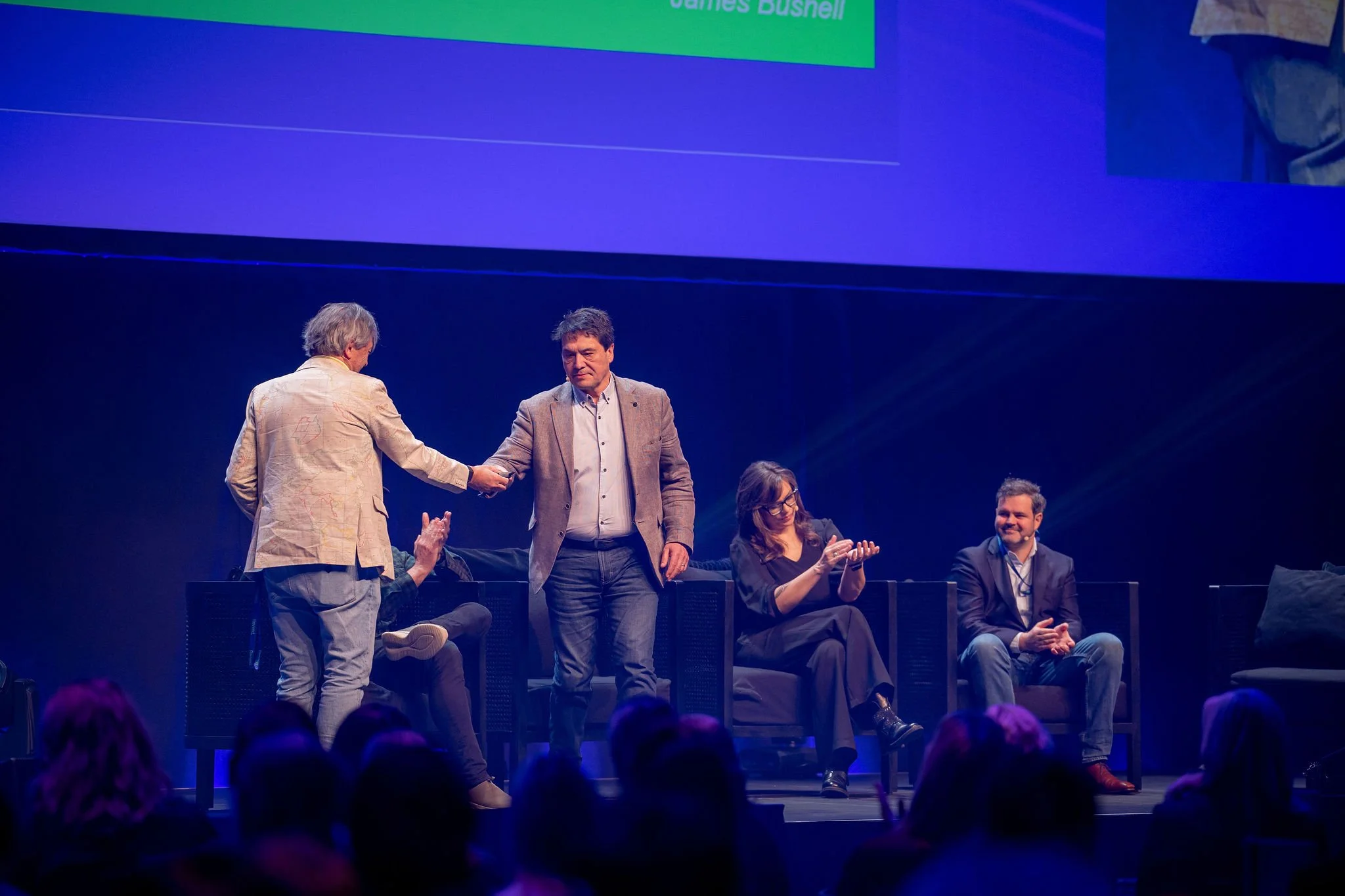 A group of people on stage at Revved, with some seated and others standing. The background includes a large screen with text, and the audience is visible in the foreground.
