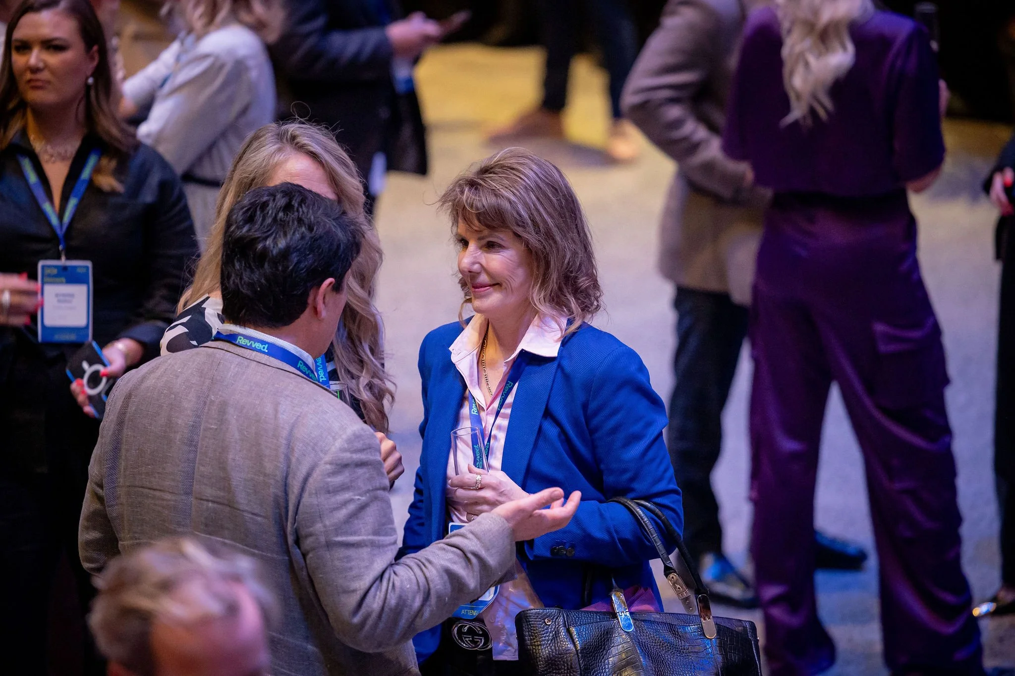 A group of people at a professional conference or event, engaged in conversation. Women are wearing conference badges; one woman in a blue blazer is smiling while talking to a man, with others in the background.