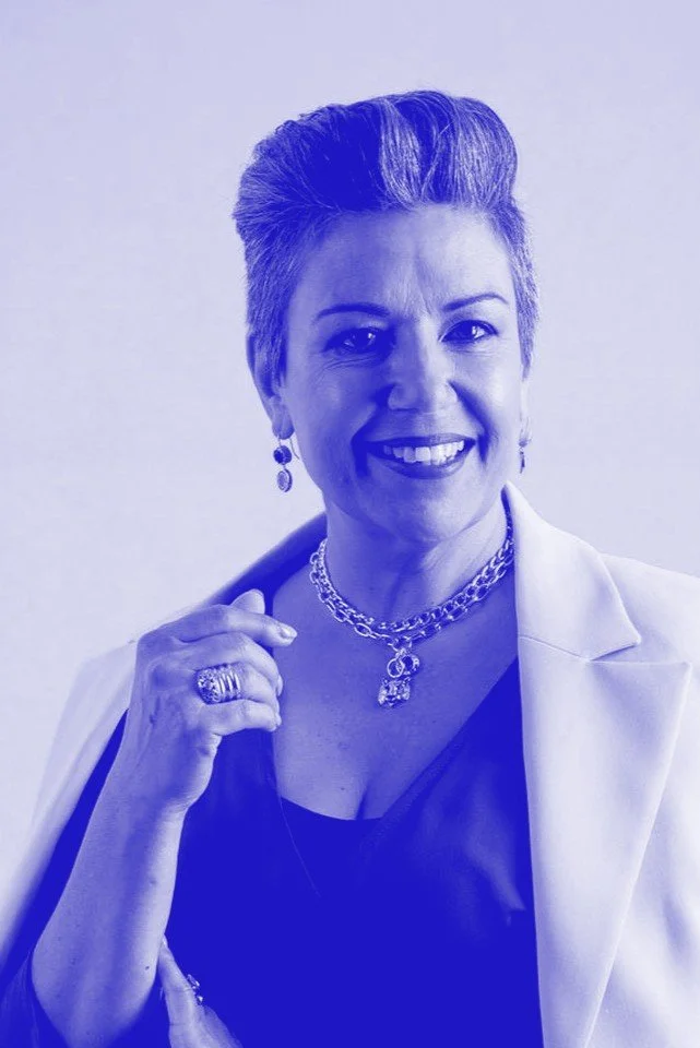 A smiling woman with short hair wearing a white blazer, black top, and multiple pieces of jewelry including rings, earrings, and layered necklaces, standing against a plain background.