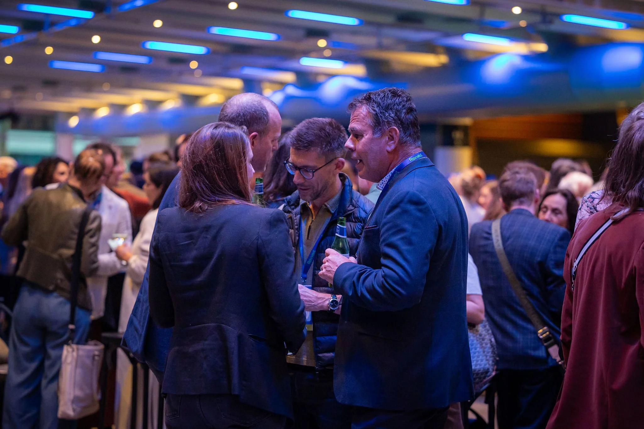 A group of people at a social event, engaged in conversation, in a venue with blue lighting and modern design elements.