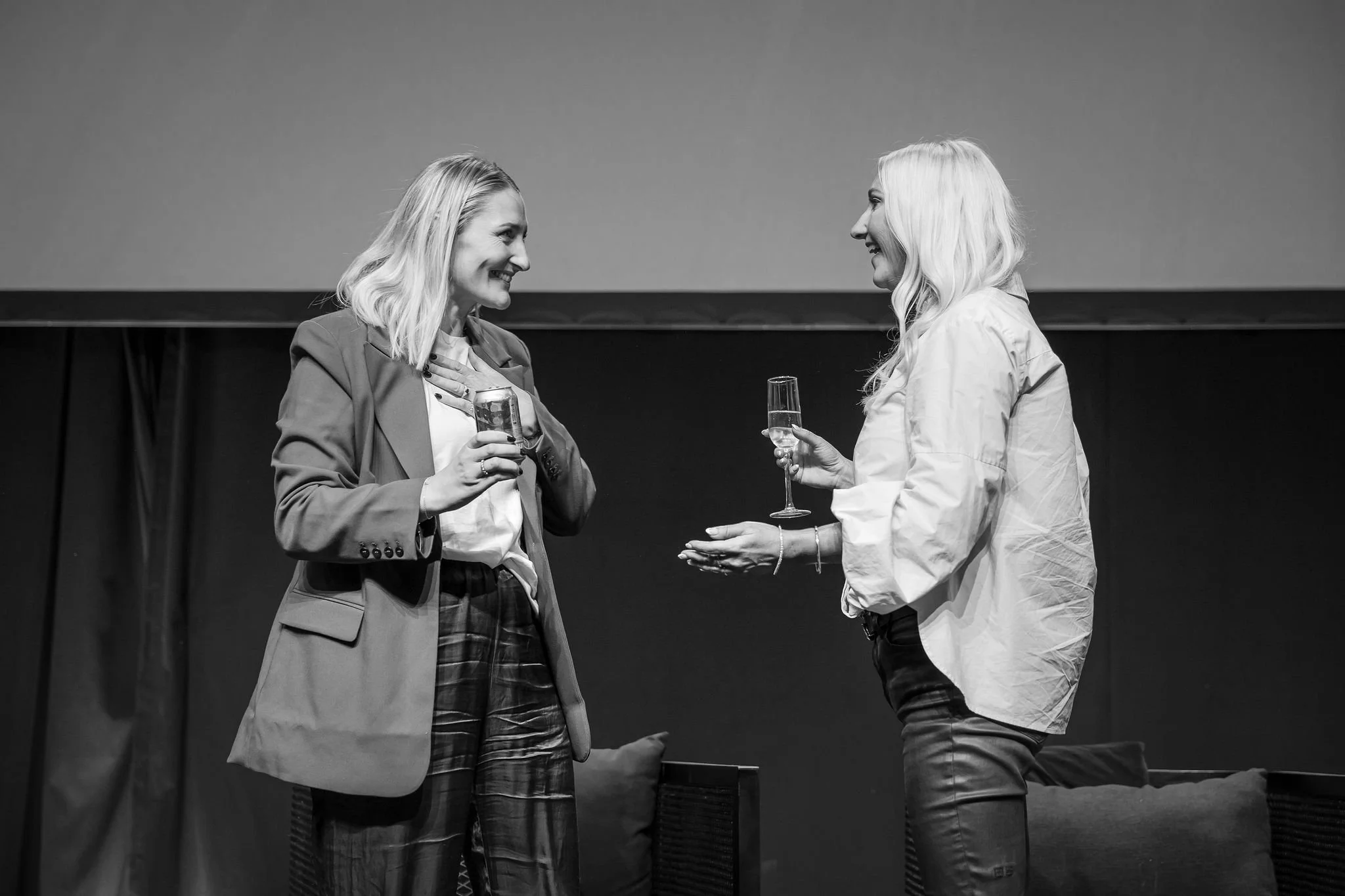 Two smiling women indoors at an event, one holding a drink, the other with a hand over her chest, engaging in conversation.