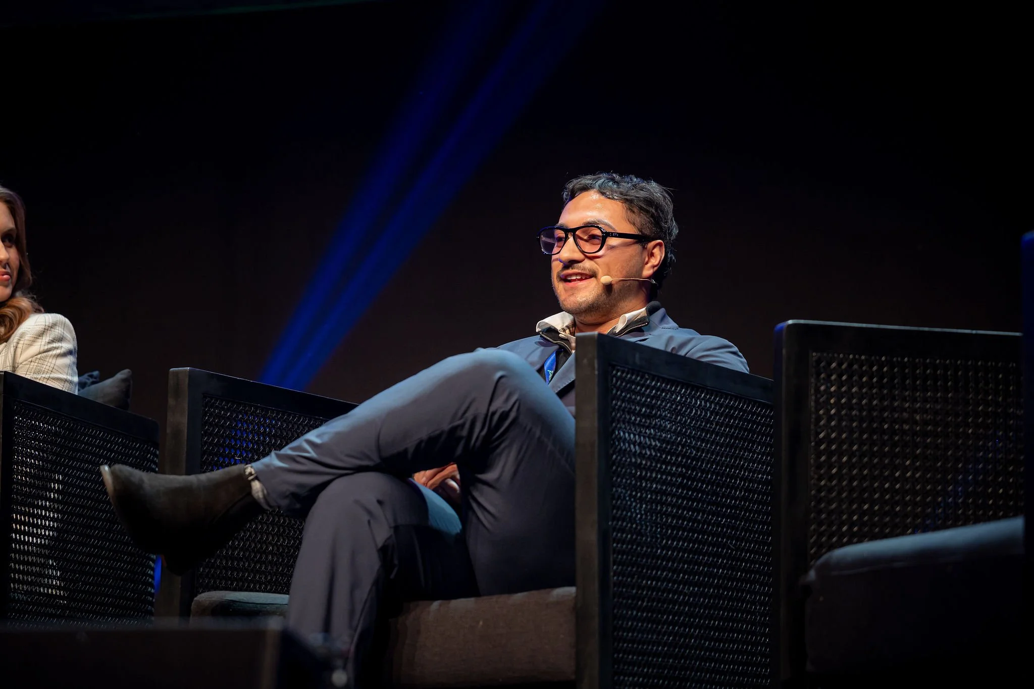 A man with glasses and a beard, wearing a dark jacket, sitting on a panel at a conference or event. Part of a woman with long, wavy hair is visible on the left side. The background is dark with blue light streaks.