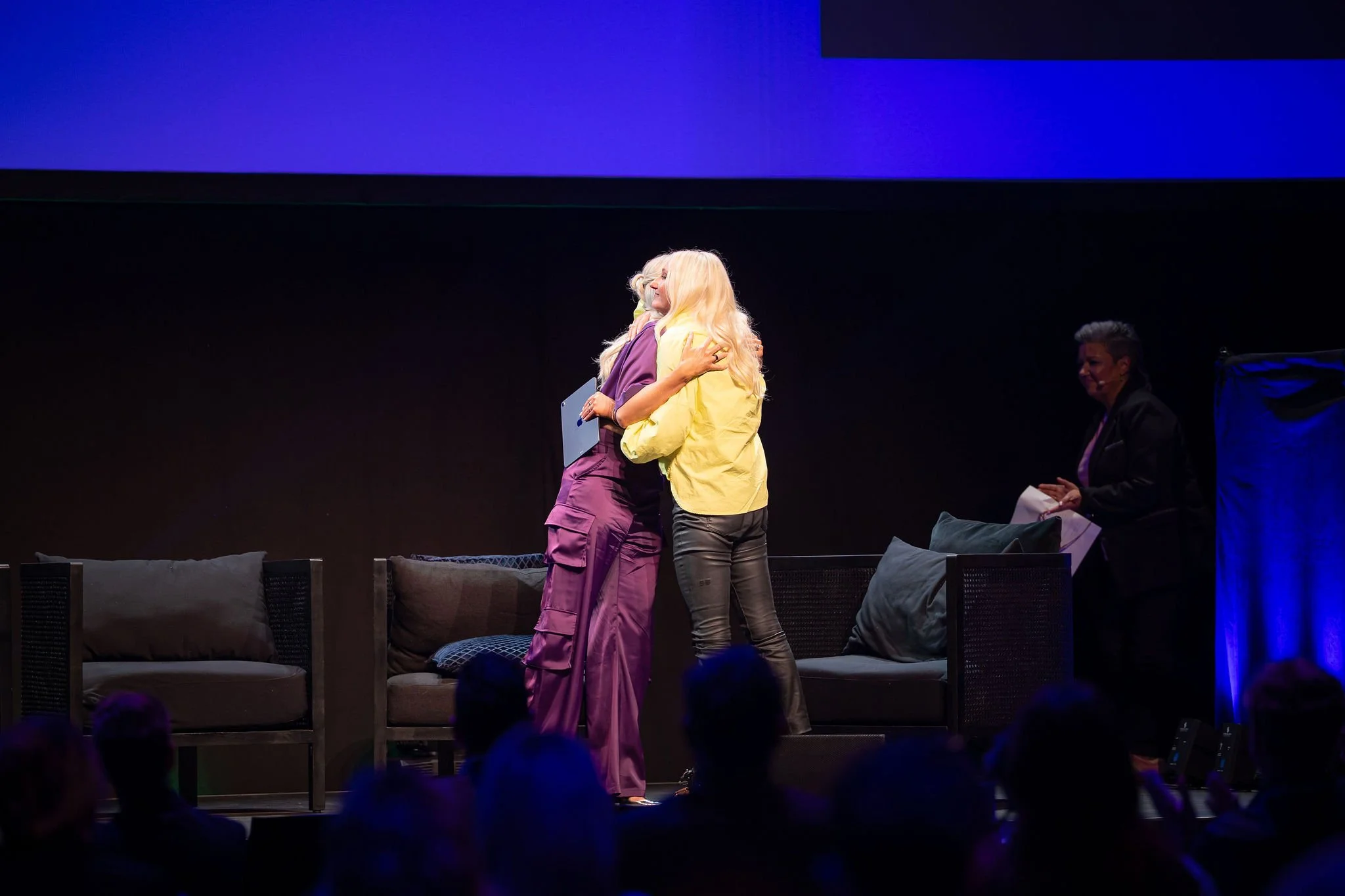 Two women are hugging on stage during an event, with a woman seated on the right holding papers and wearing a dark blazer, and a dark-colored sofa with cushions is visible in front of them.