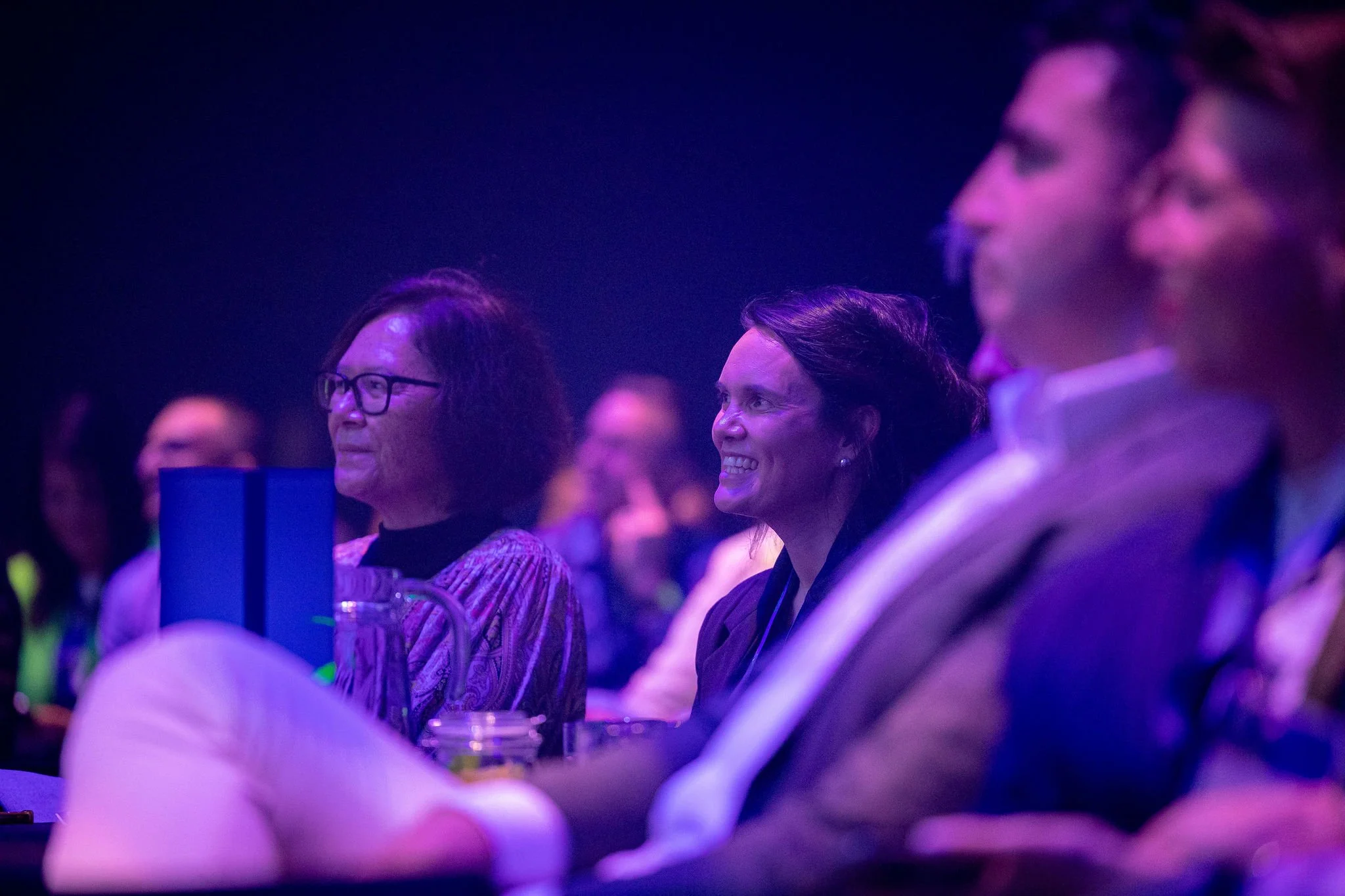 Revved audience members watching a stage performance in a dark, purple-lit theater, smiling and focused.
